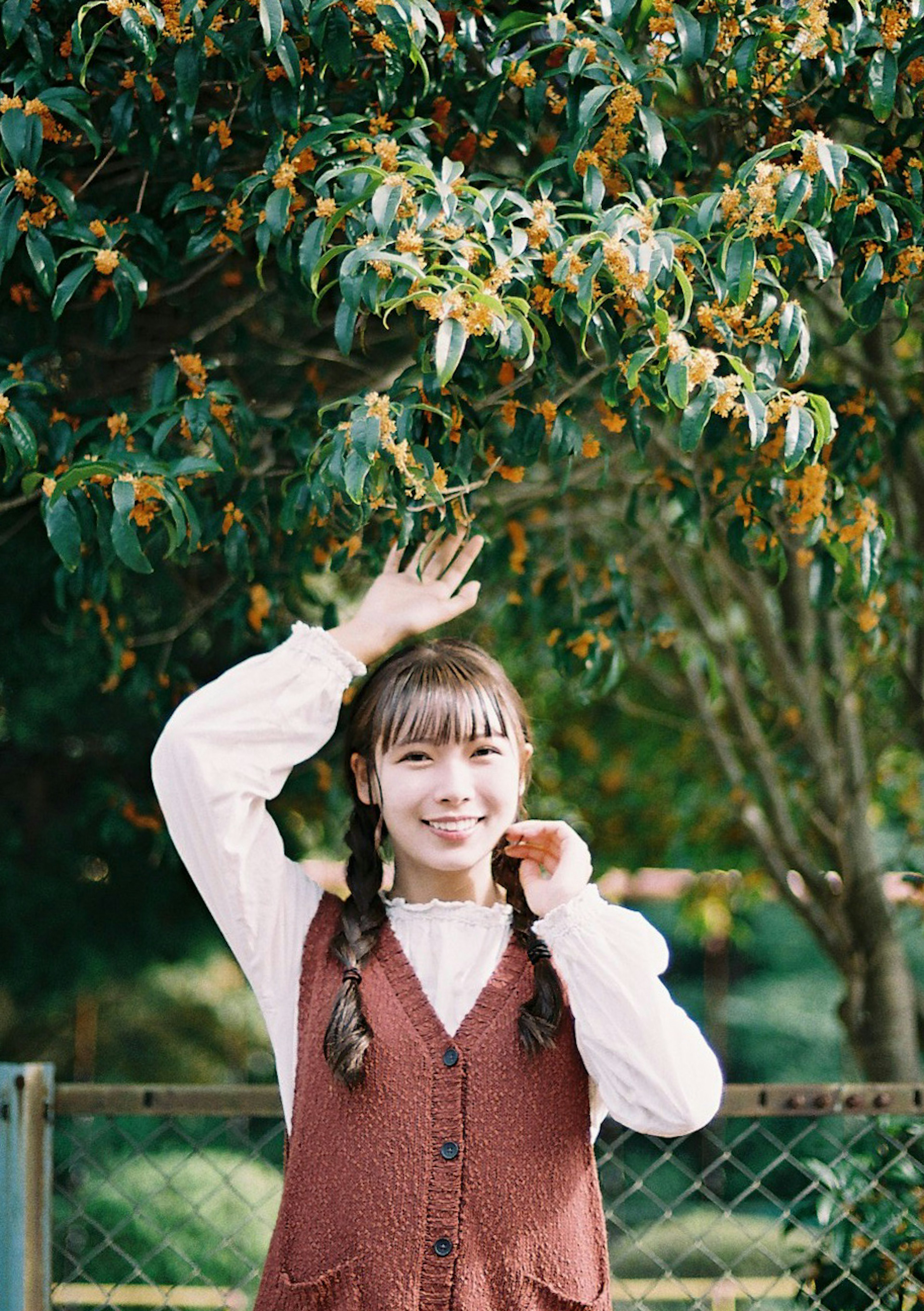 Fille posant devant un arbre vert portant un gilet marron et une chemise blanche