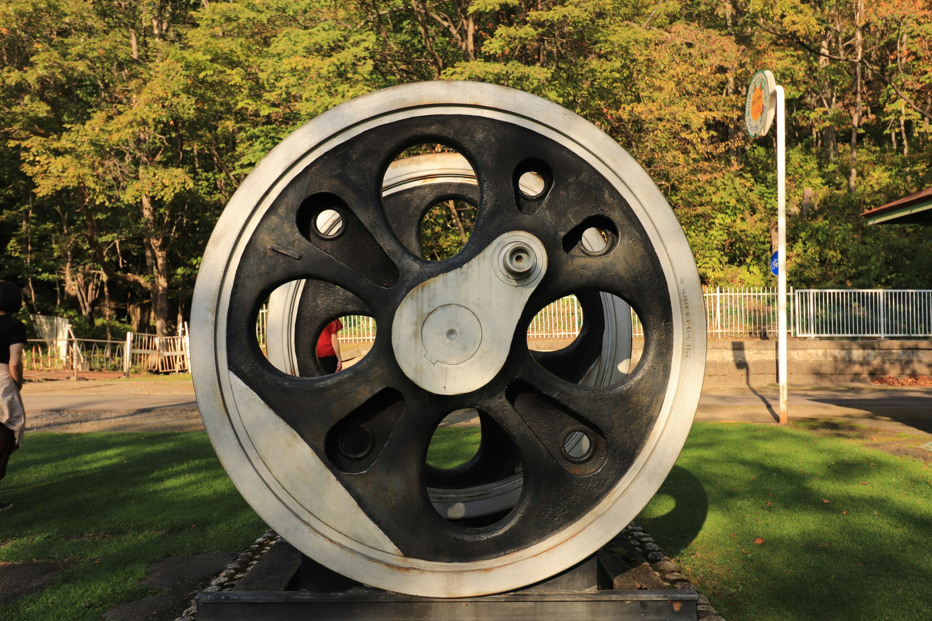 Large iron wheel sculpture in a park black and silver color scheme green trees in the background