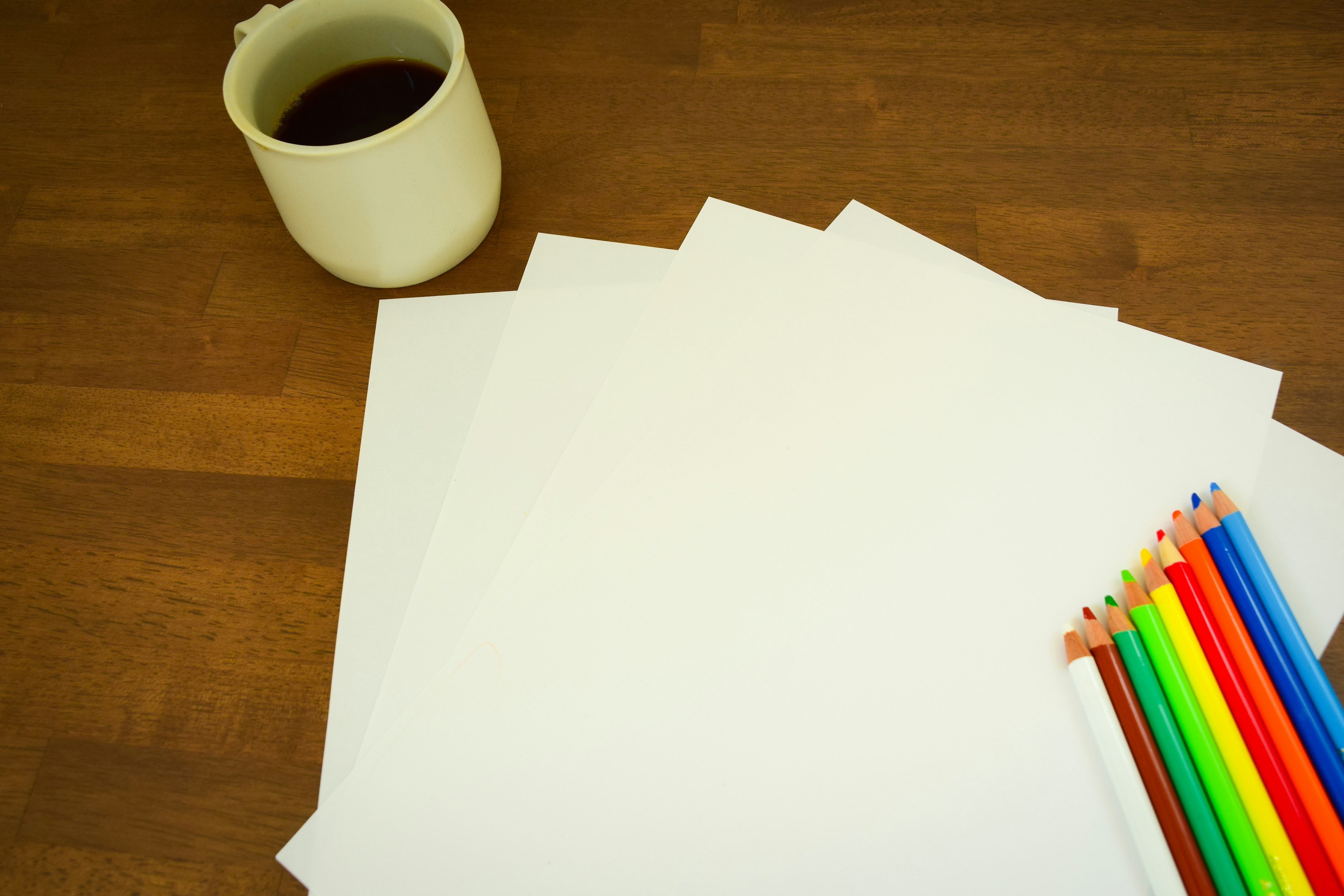 Una mesa con hojas de papel en blanco y lápices de colores junto a una taza de café