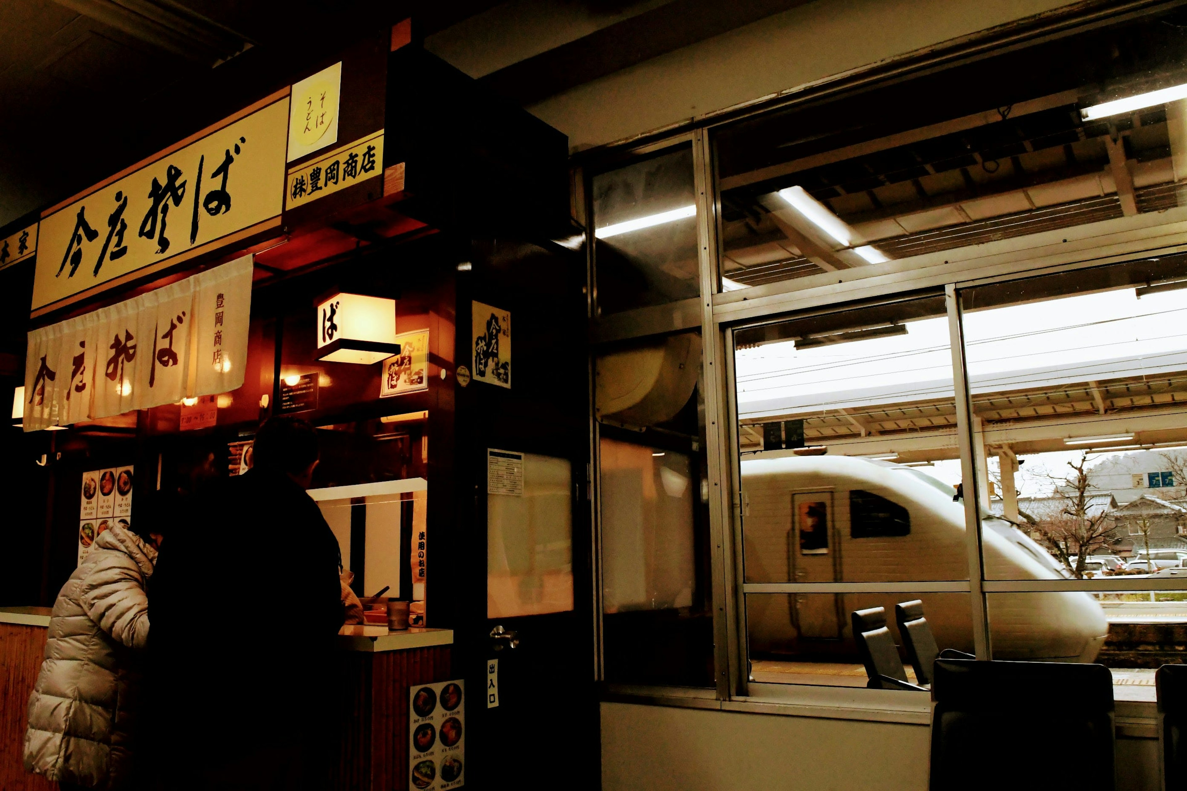 Un ristorante alla stazione con un cliente che ordina soba e un Shinkansen visibile all'esterno