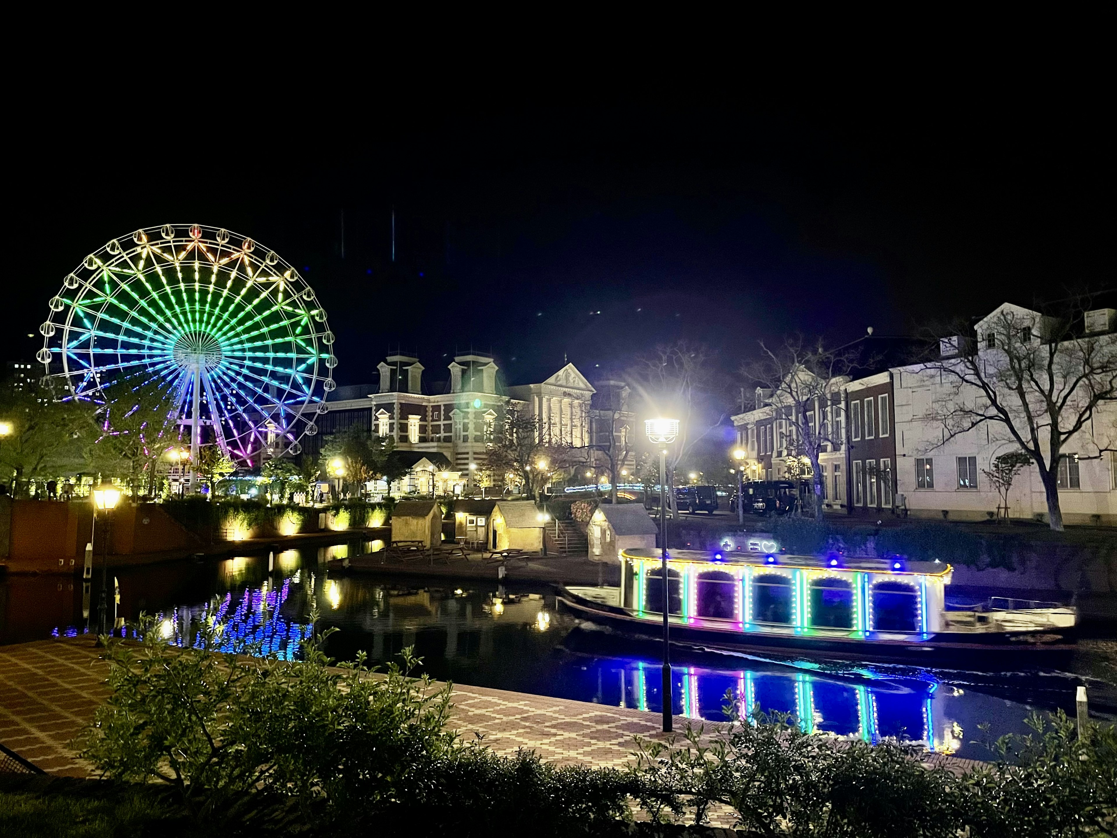 Nachtansicht mit einem bunten Riesenrad und einem beleuchteten Boot auf dem Fluss