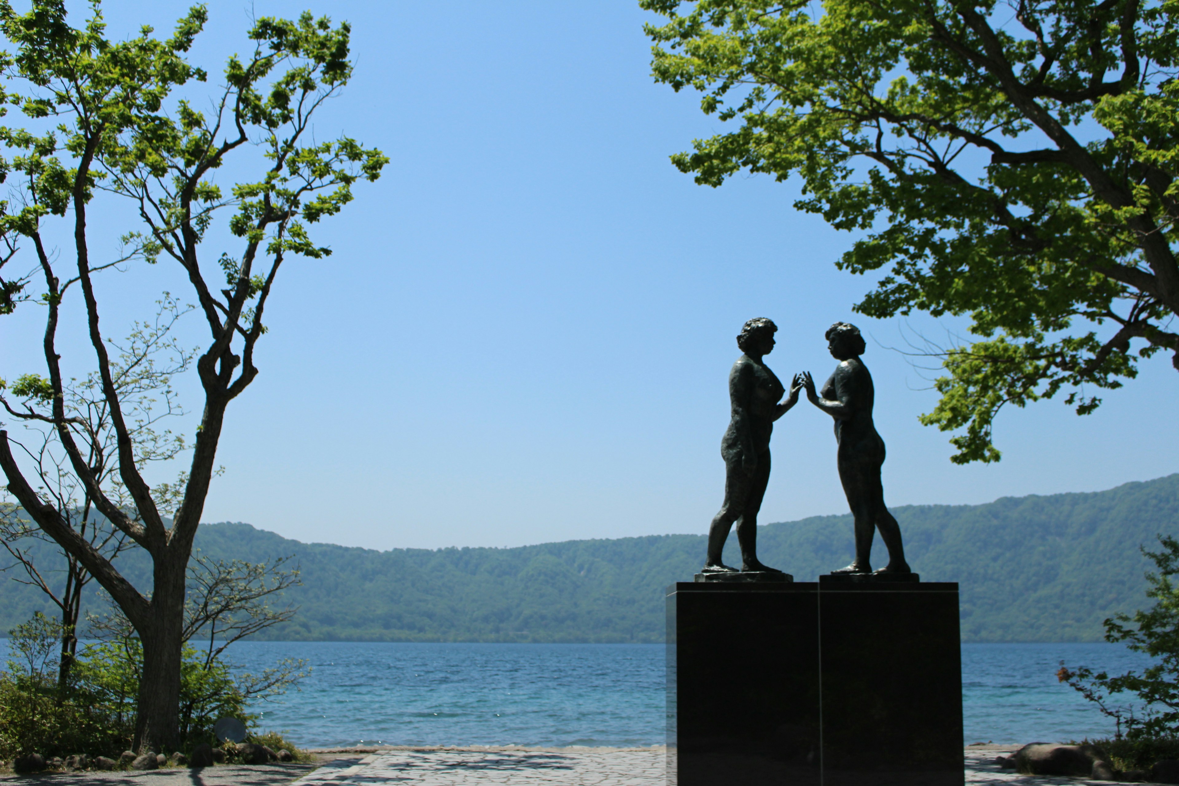 Escultura de dos figuras tomándose de las manos junto a un lago con cielo azul claro