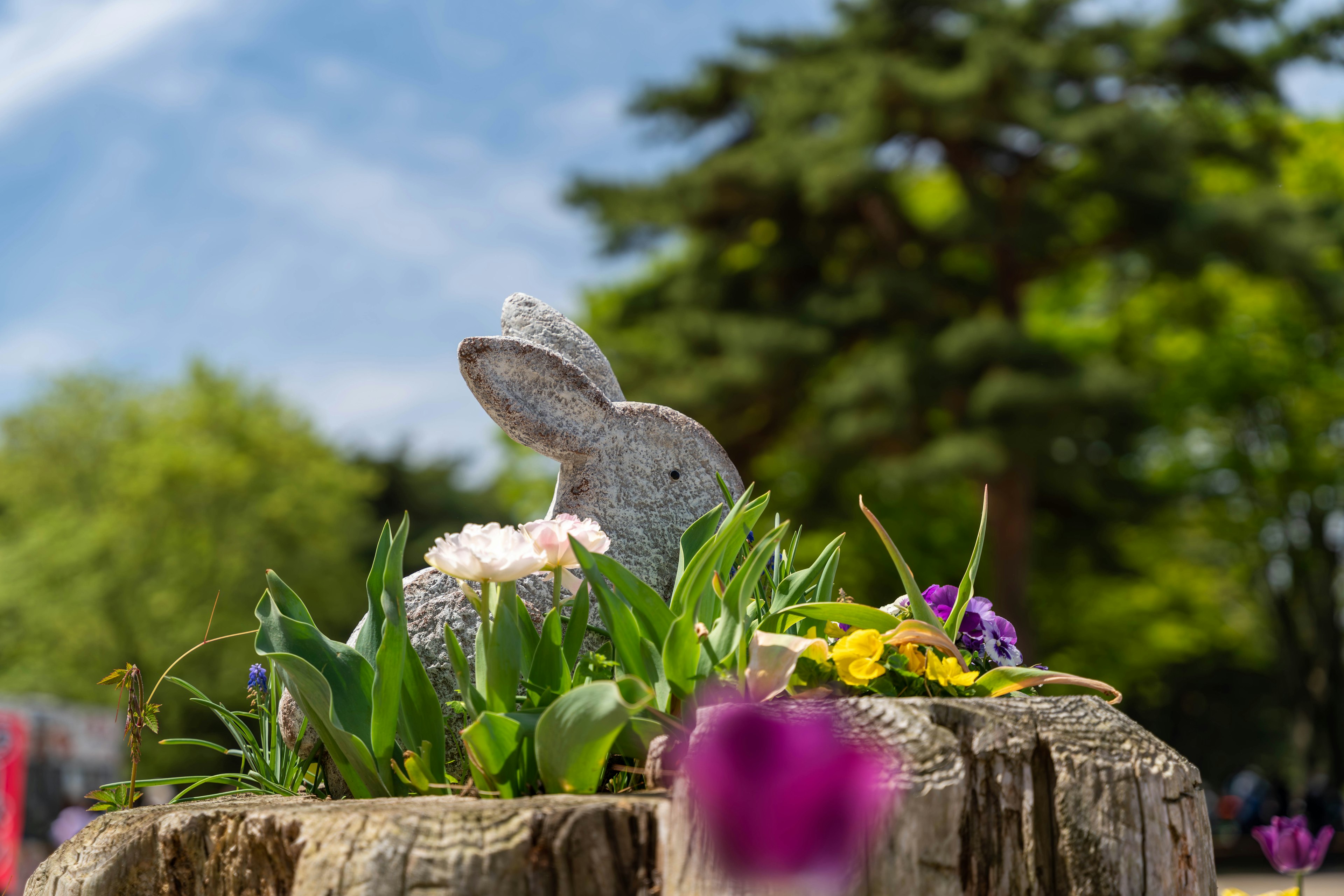 A rabbit figurine surrounded by flowers and grass on a tree stump