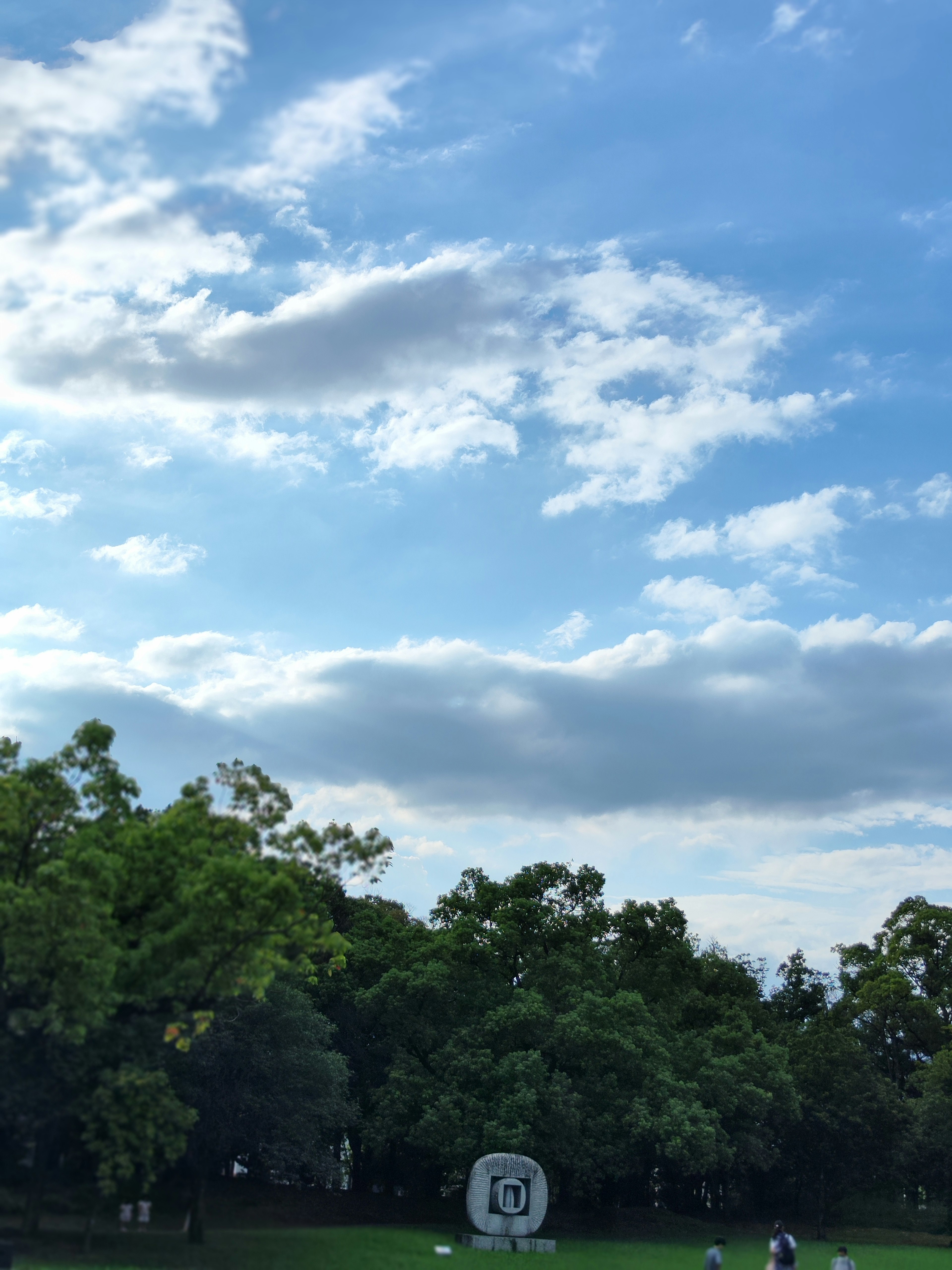 Parkszene mit blauem Himmel und weißen Wolken grünen Bäumen und einer Steinskulptur