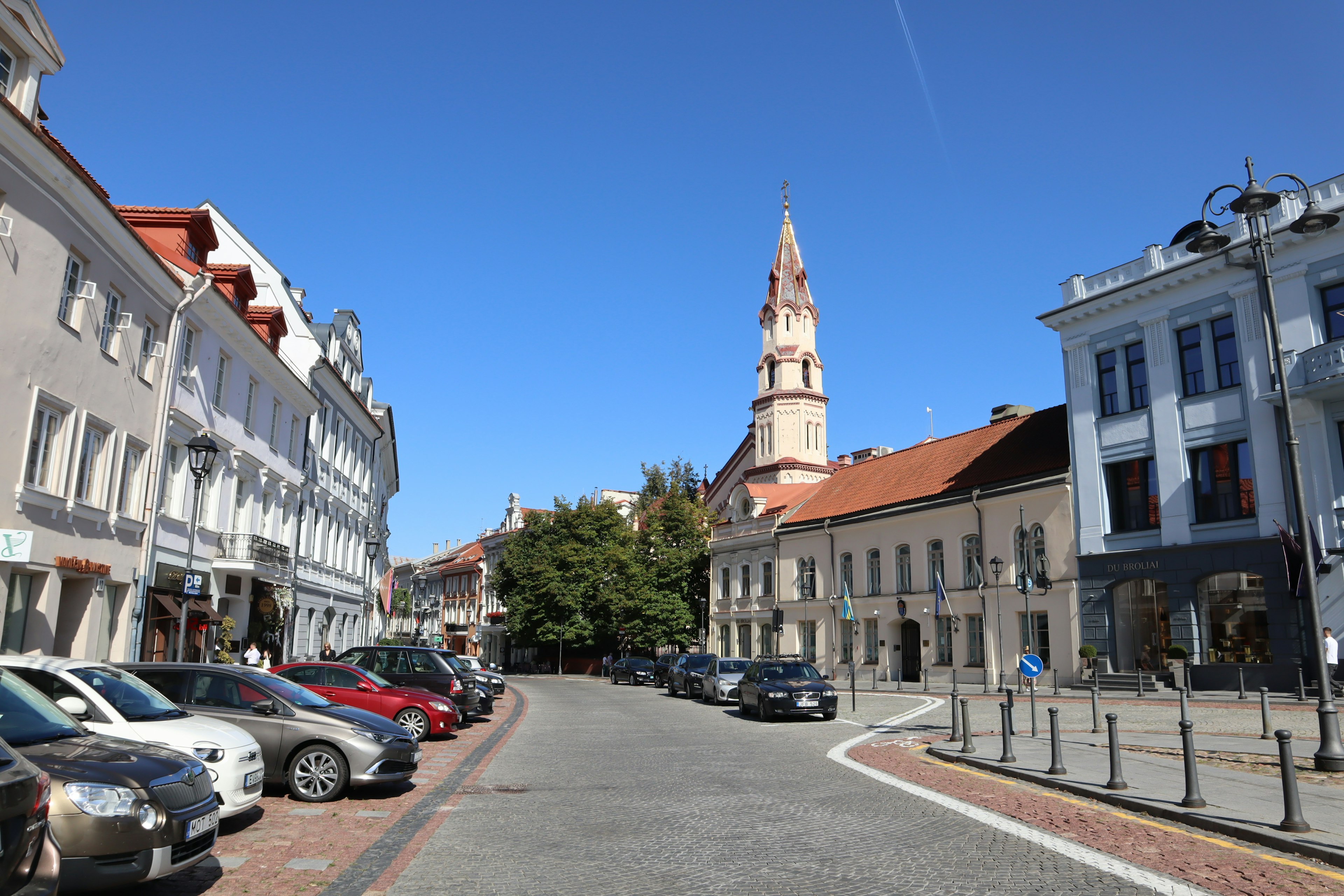 Strada cittadina con auto parcheggiate e torre di chiesa sotto un cielo blu