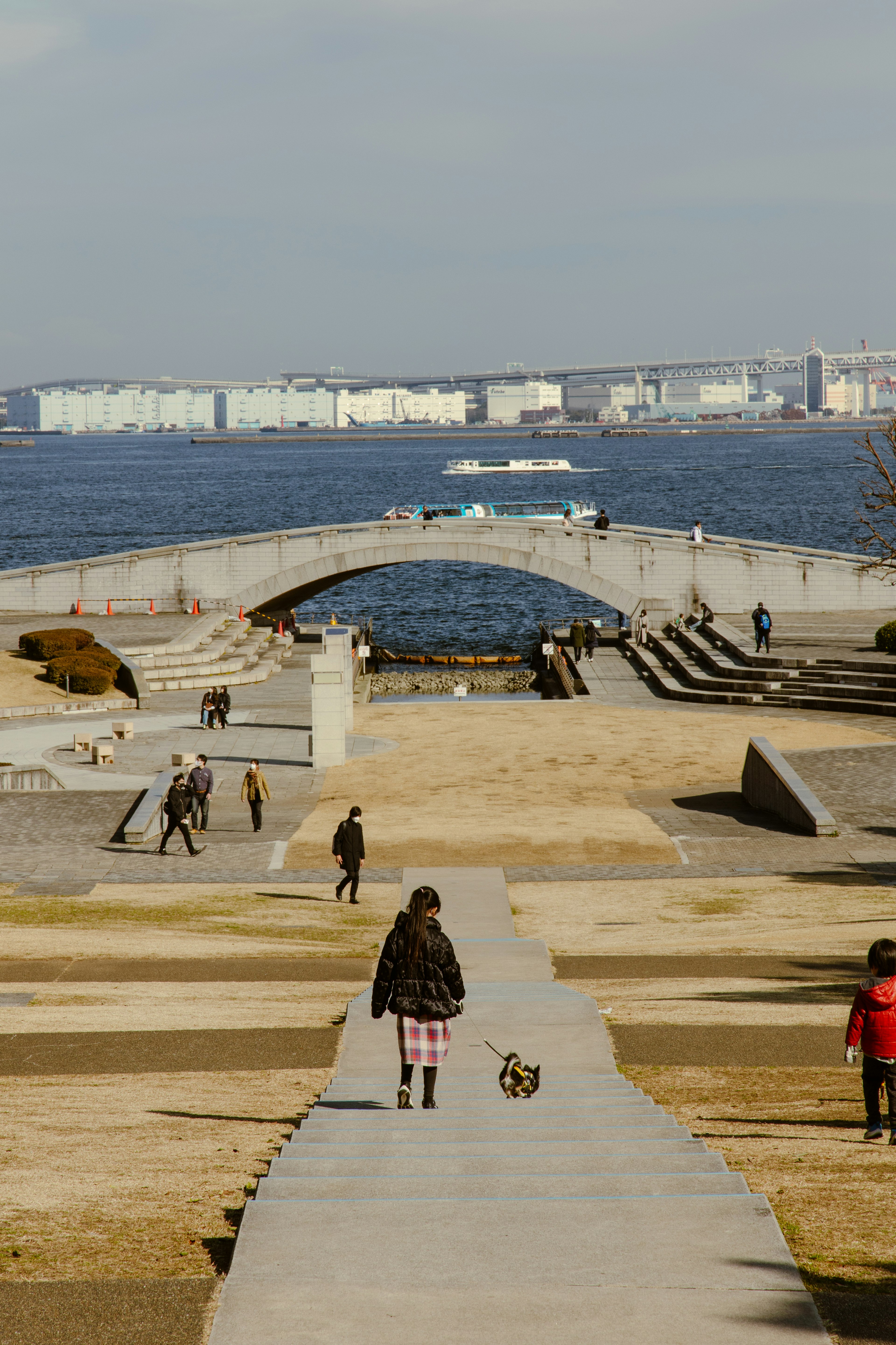 People walking in a park near a river with a bridge in the background