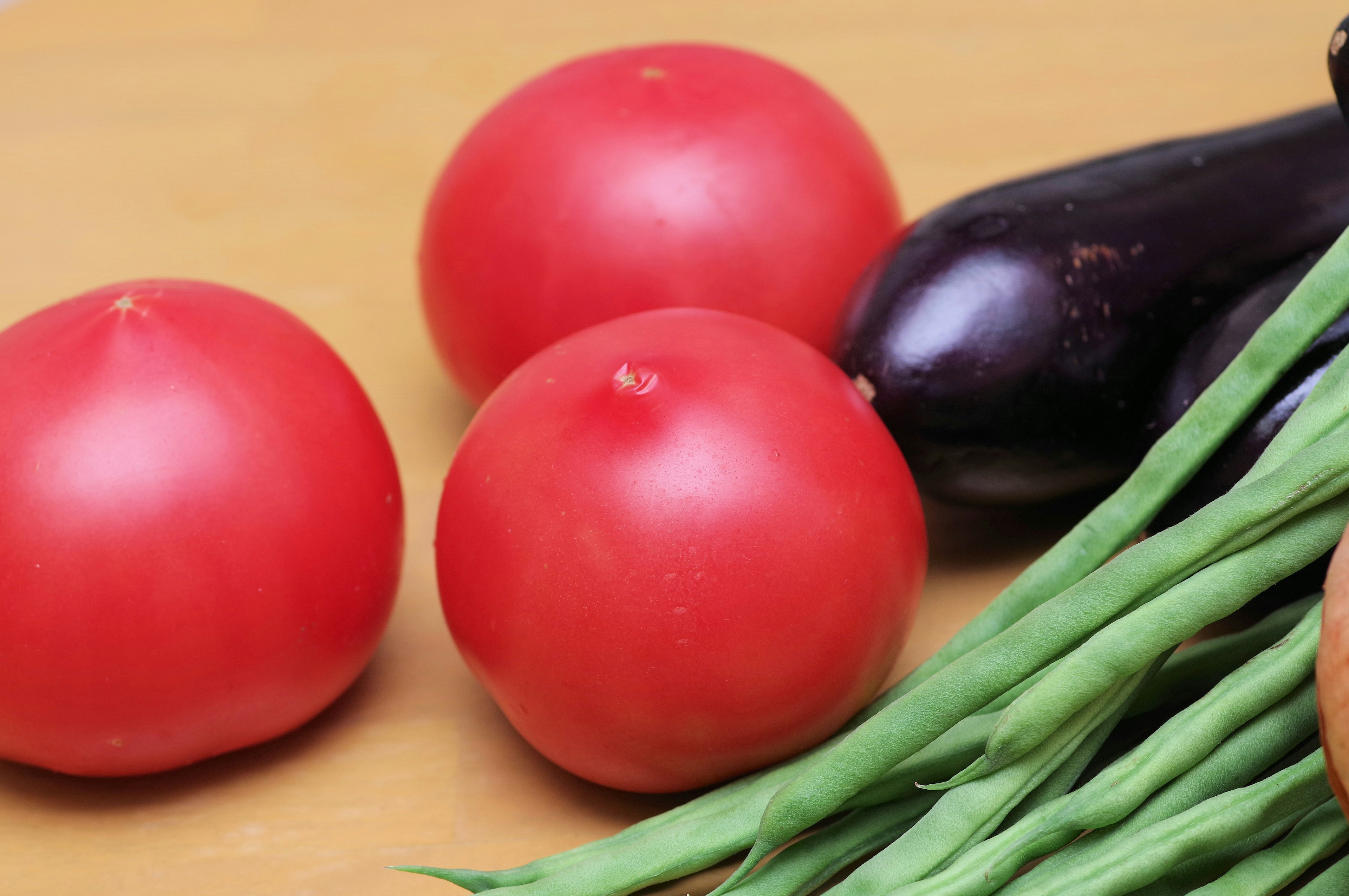 Three red tomatoes with an eggplant and green beans on a wooden surface