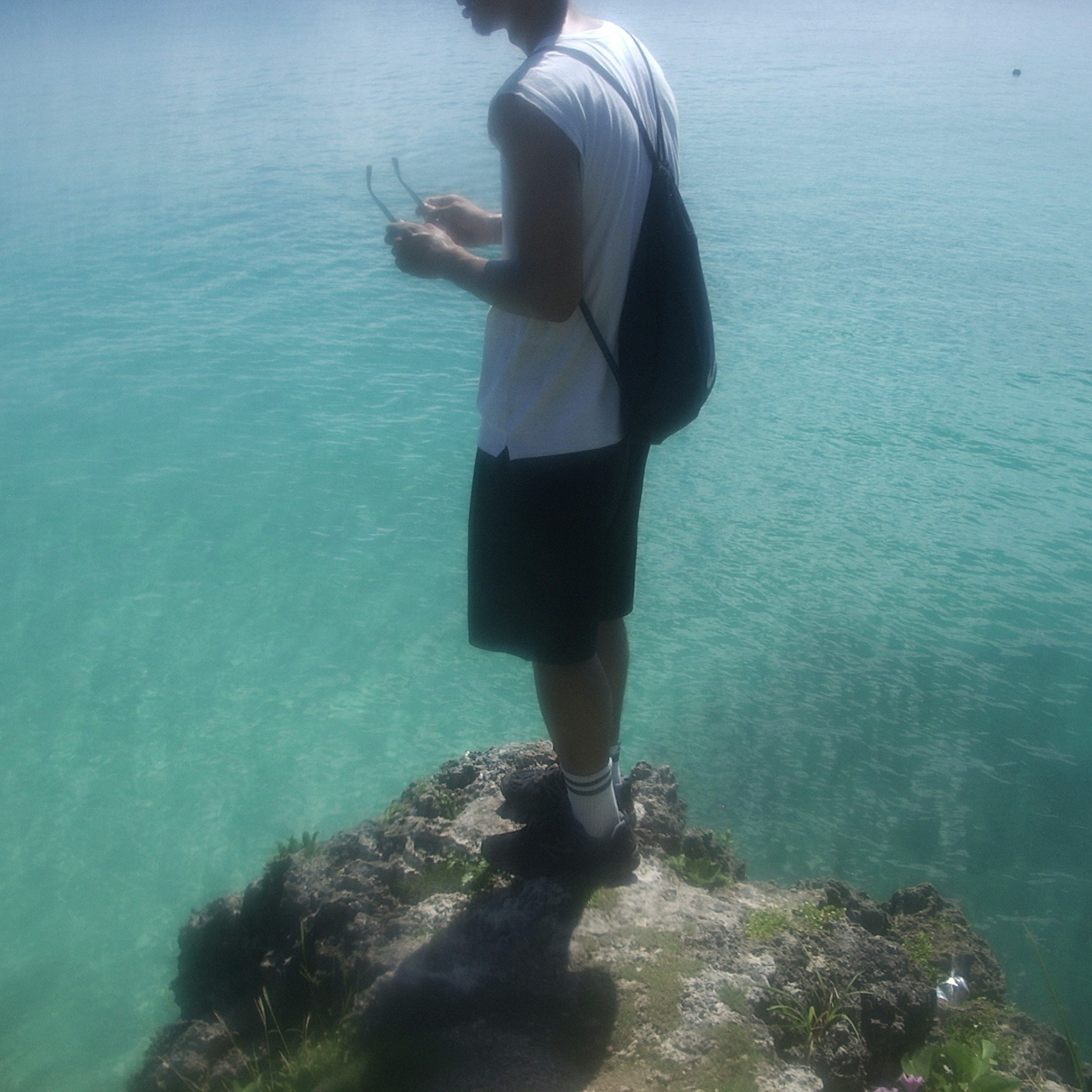 Junger Mensch steht auf einem Felsen am Meer trägt ein weißes T-Shirt und schwarze Shorts mit einem Rucksack