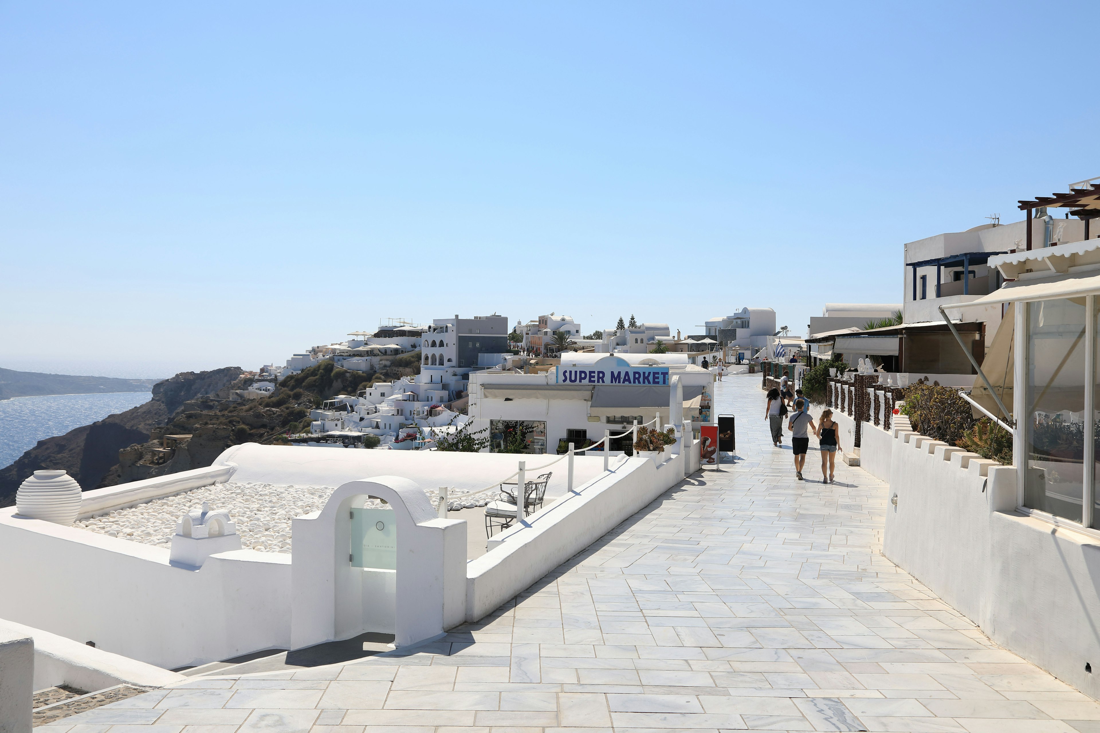 Personas caminando por un camino costero en Santorini bajo un cielo azul claro con edificios blancos