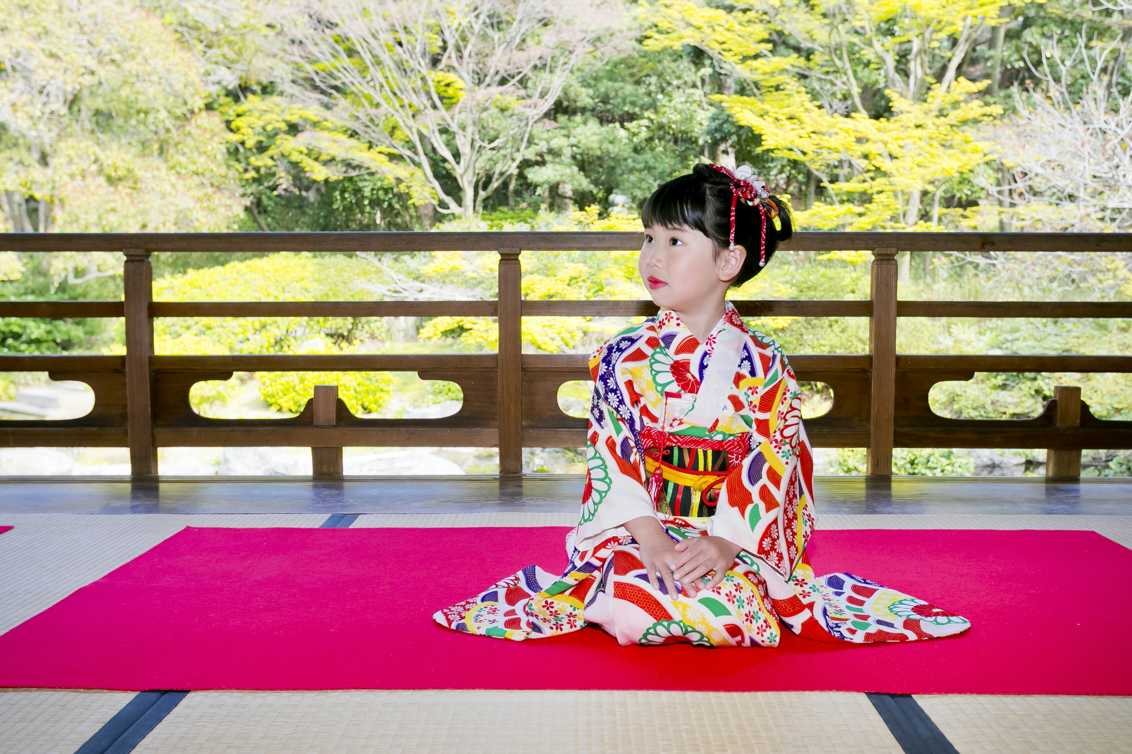 Gadis berpakaian kimono cantik duduk di atas karpet merah dengan latar belakang hijau yang rimbun