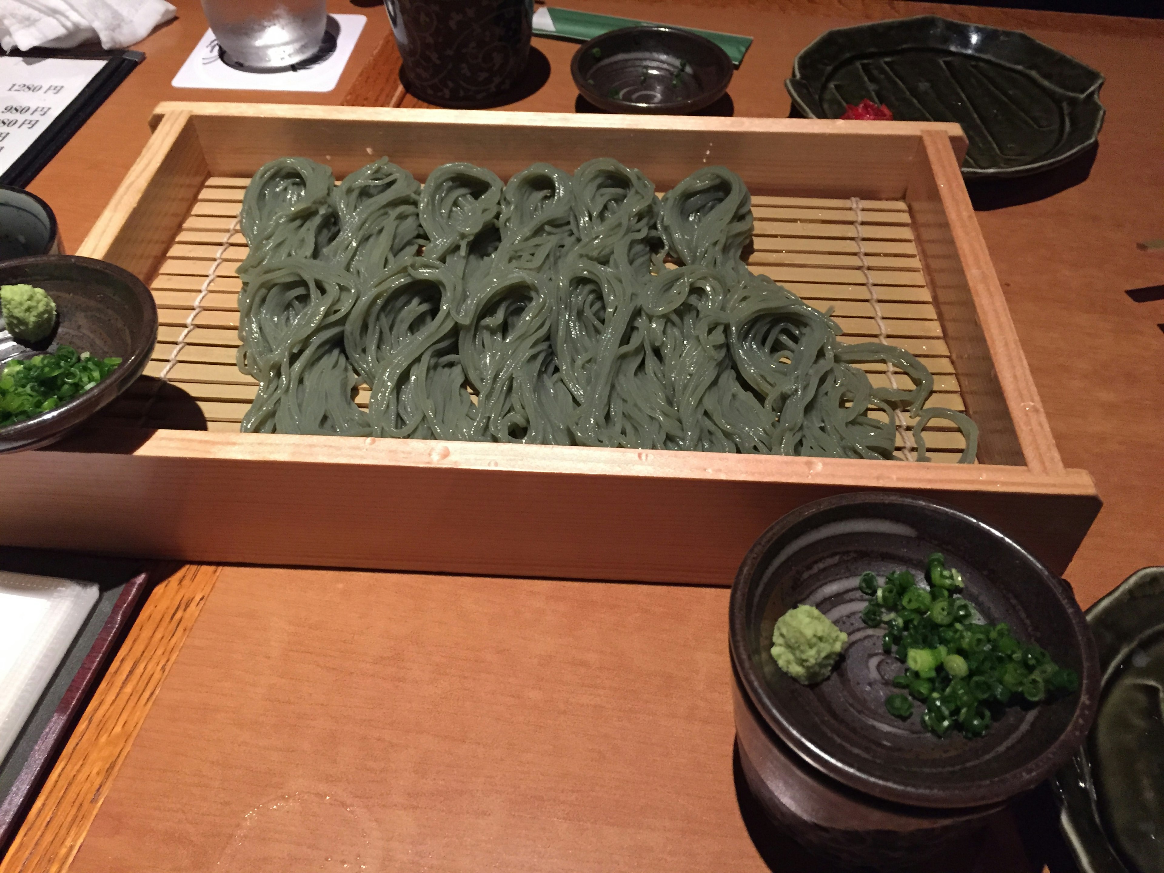 A wooden tray with green soba noodles served alongside condiments like wasabi and green onions