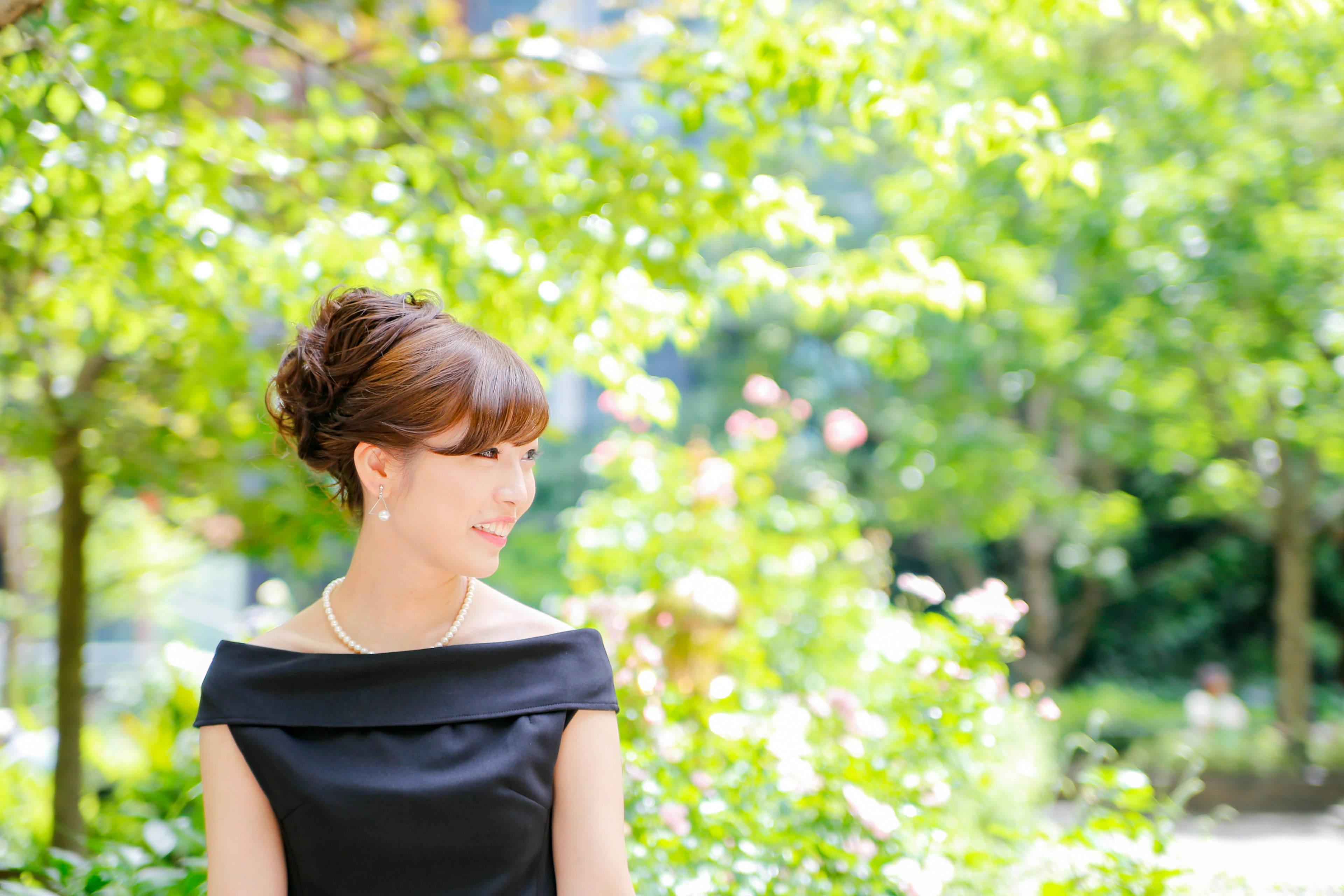 Femme souriante au milieu des arbres verts portant une robe noire et un collier de perles