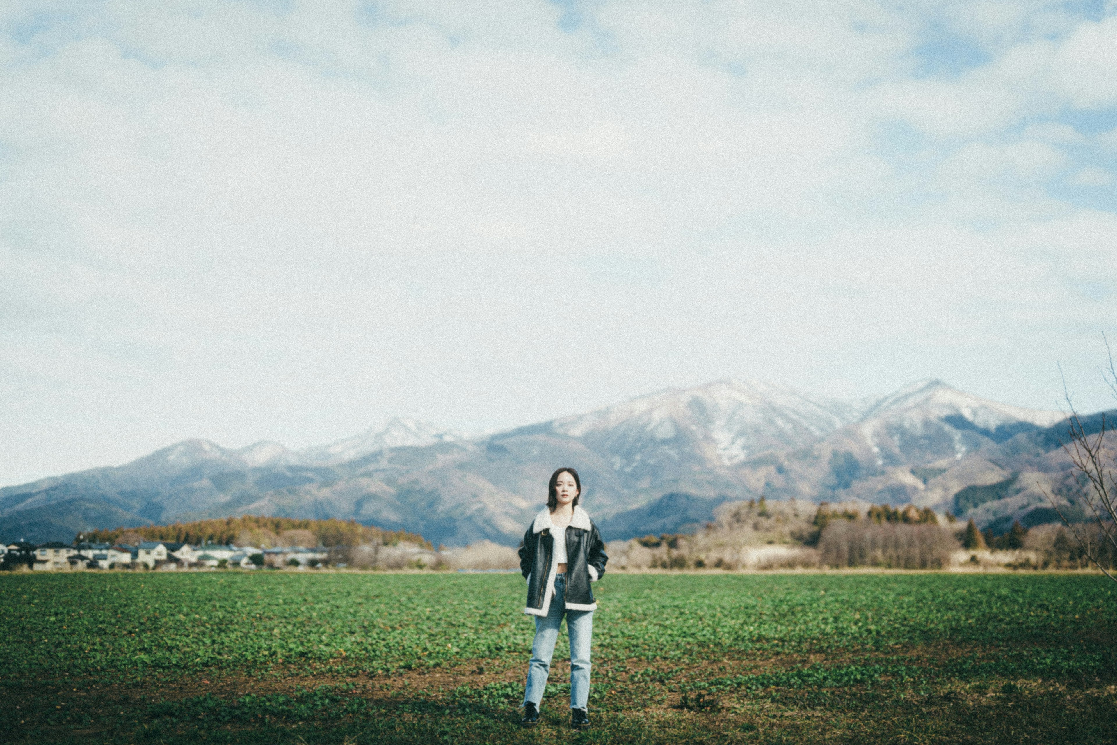 A person standing in a vast green field with mountains in the background and a cloudy sky above