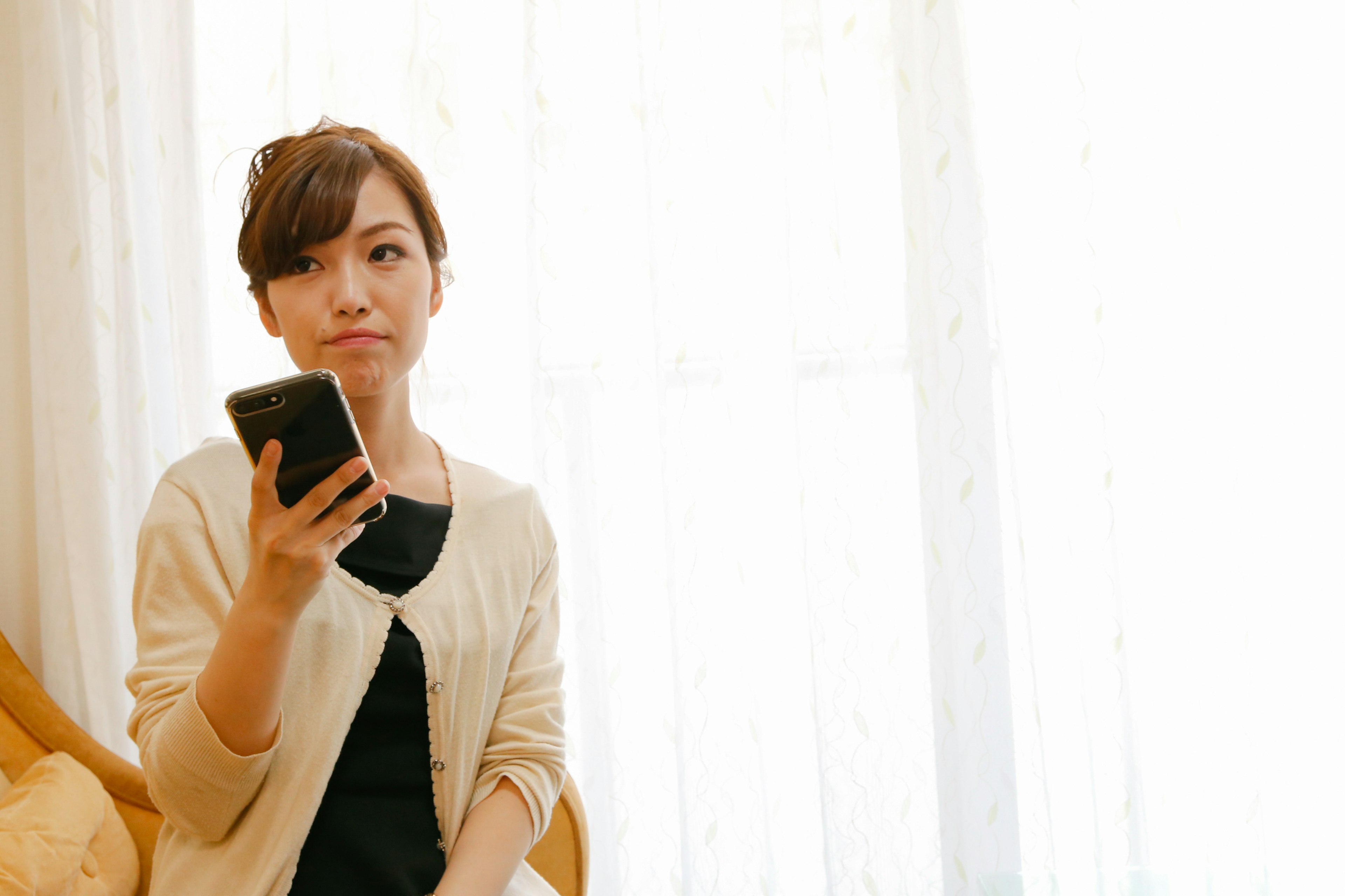 Mujer sosteniendo un teléfono inteligente sentada cerca de una ventana con aspecto pensativo