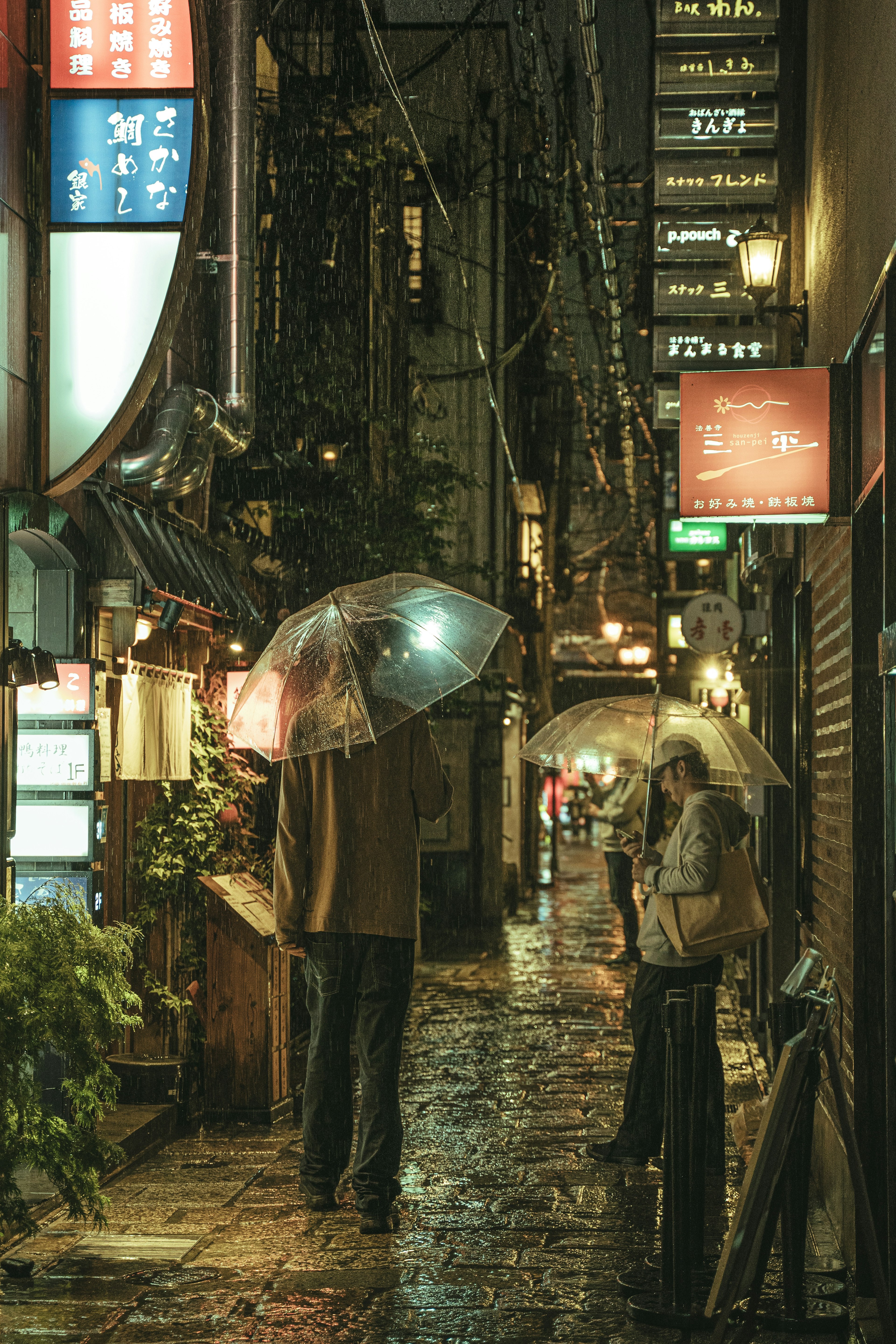 雨の中の狭い路地に傘を持つ人々の姿と明るい看板がある