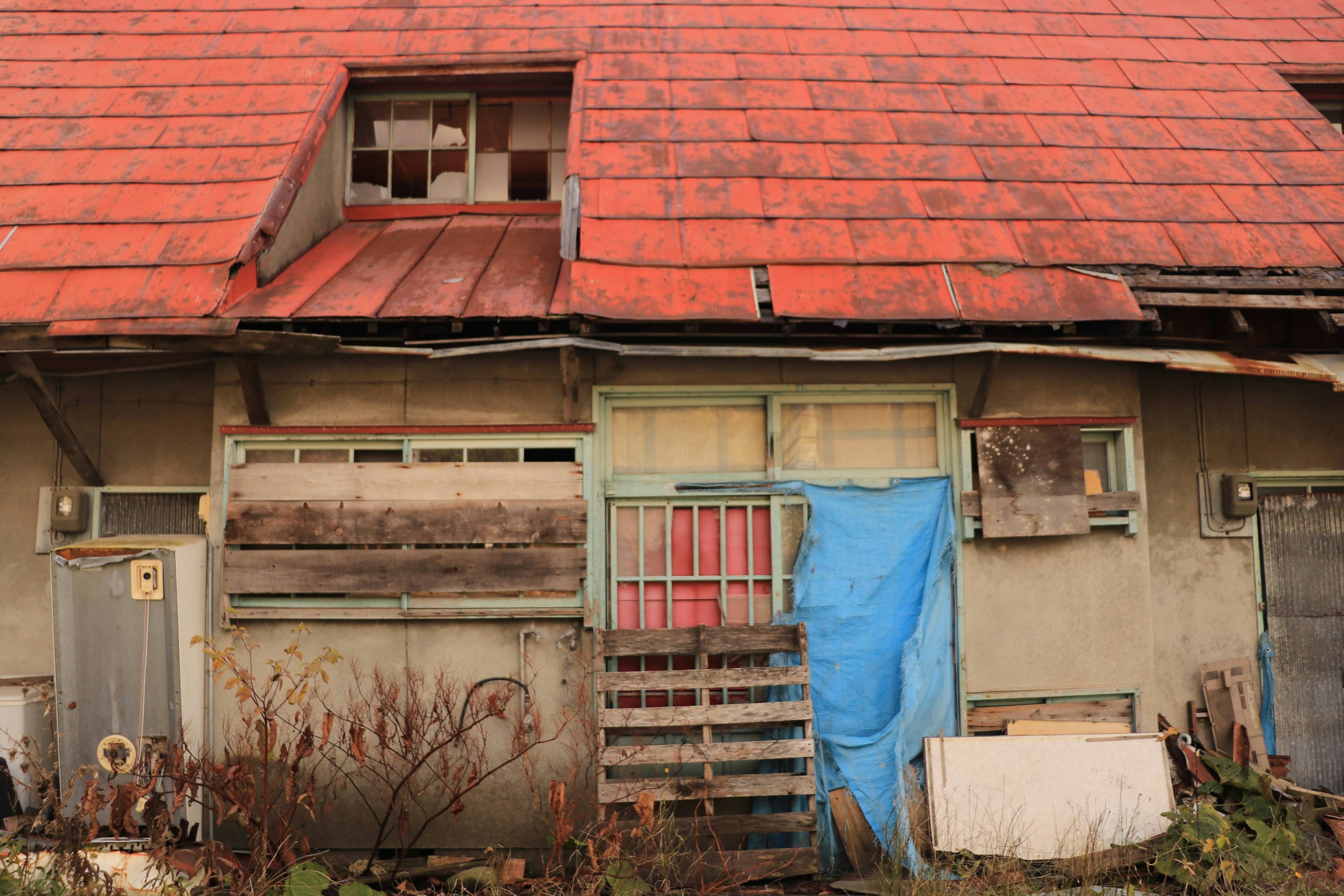 Exterior de una casa vieja con techo de tejas ventanas rotas y una puerta cubierta con tela azul