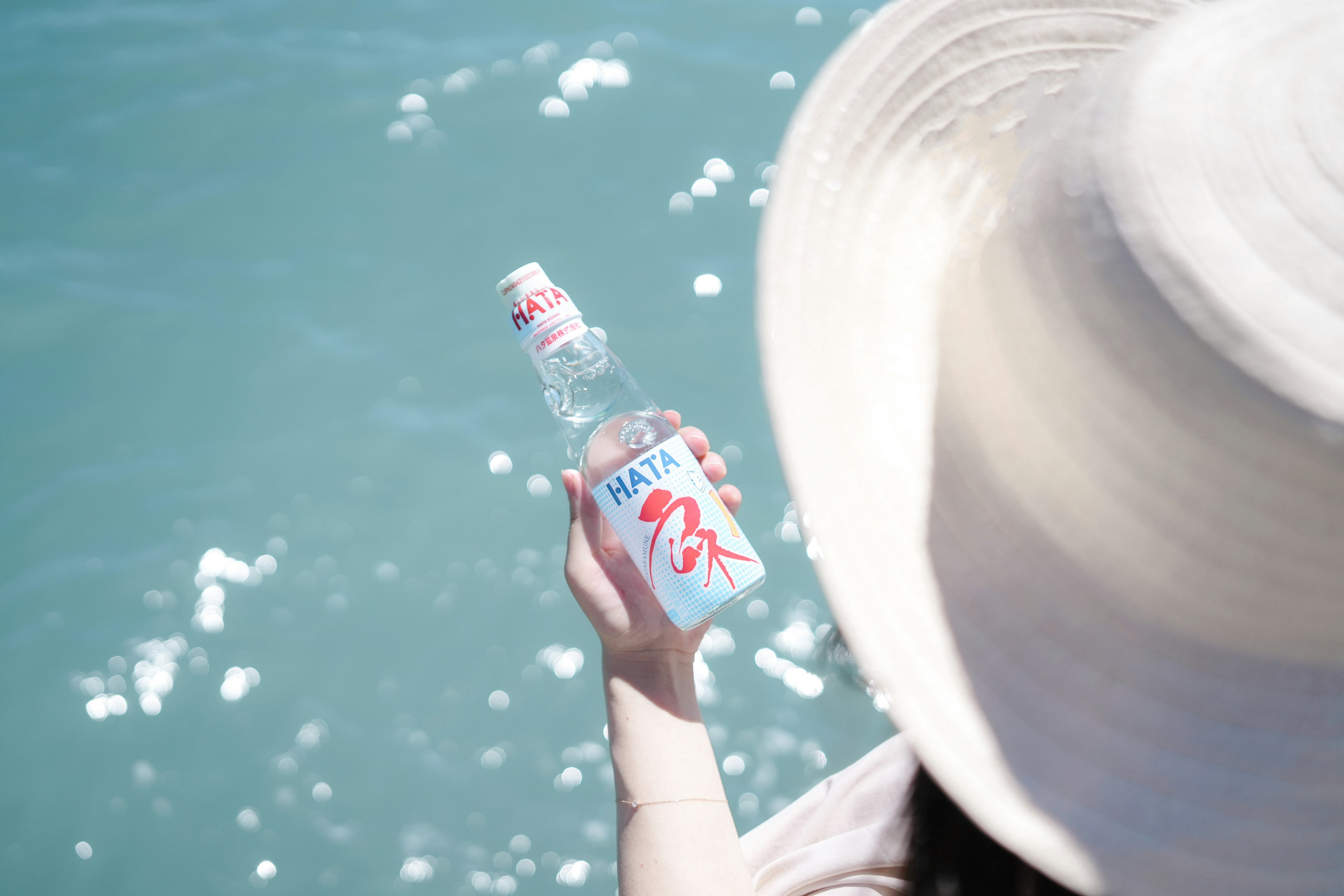 Eine Hand einer Frau, die eine Wasserflasche nahe dem blauen Meer mit einem großen Hut hält