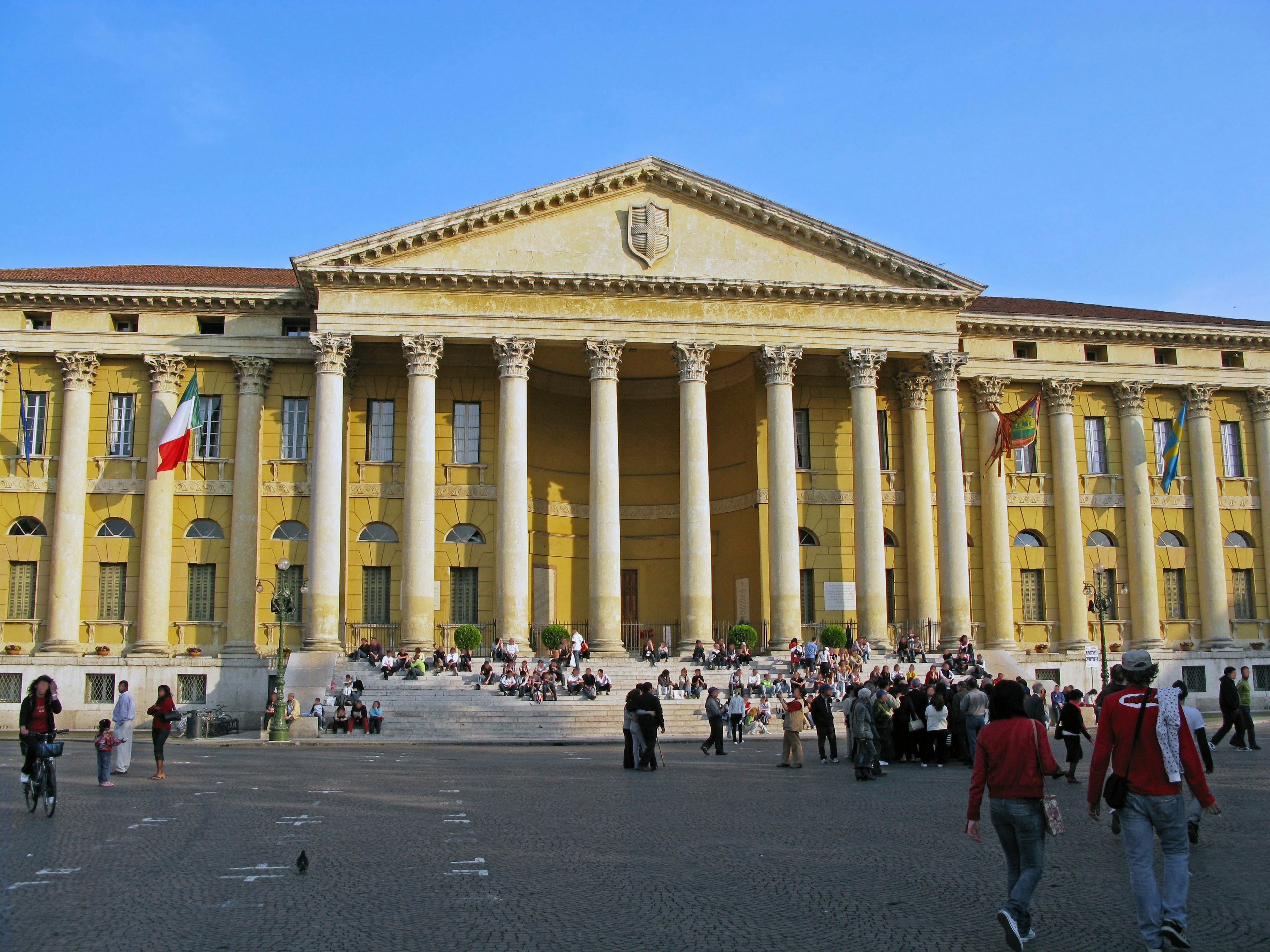 Personnes devant un grand bâtiment jaune avec des colonnes et des drapeaux