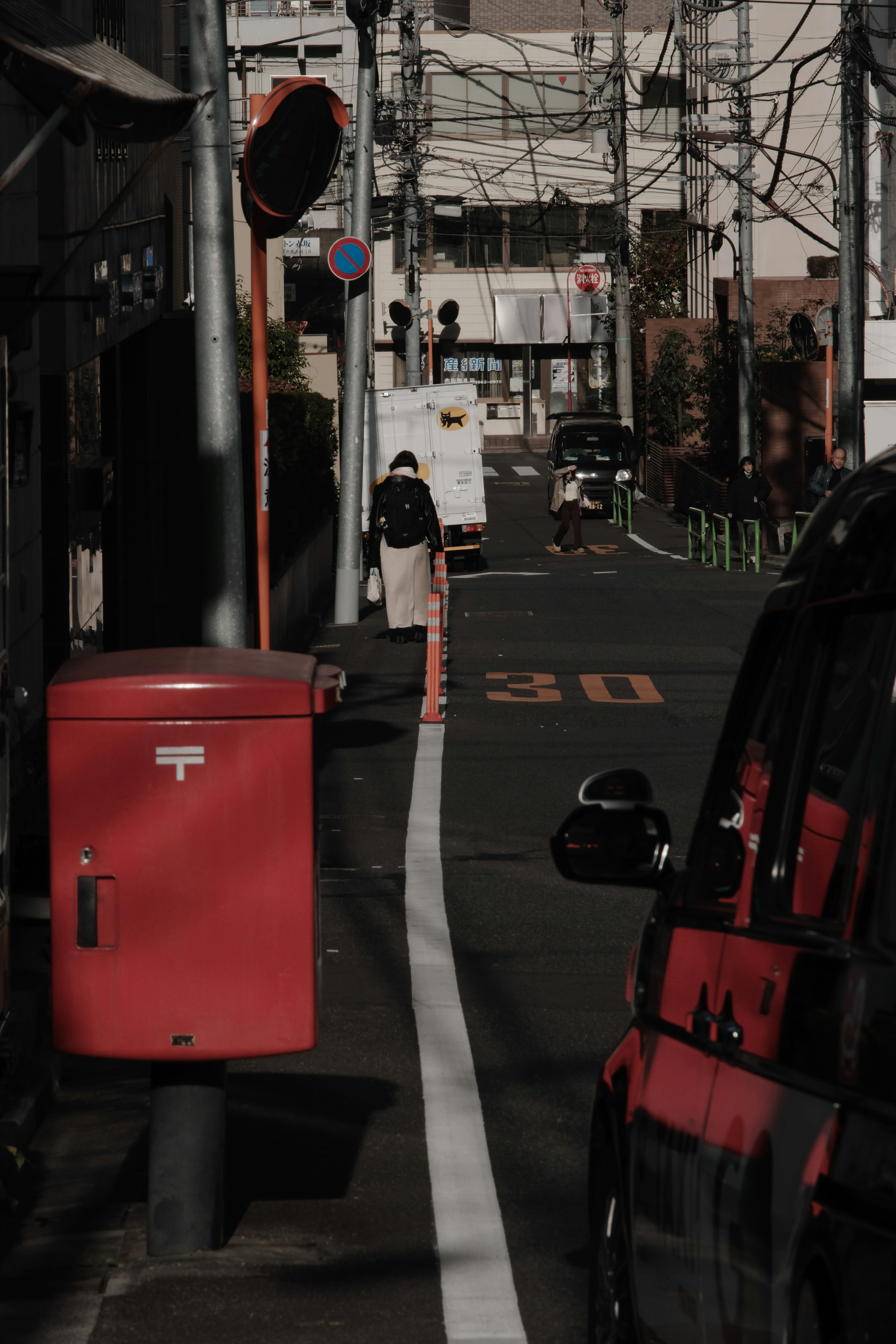 Calle estrecha con un buzón rojo y una persona caminando junto a una línea blanca