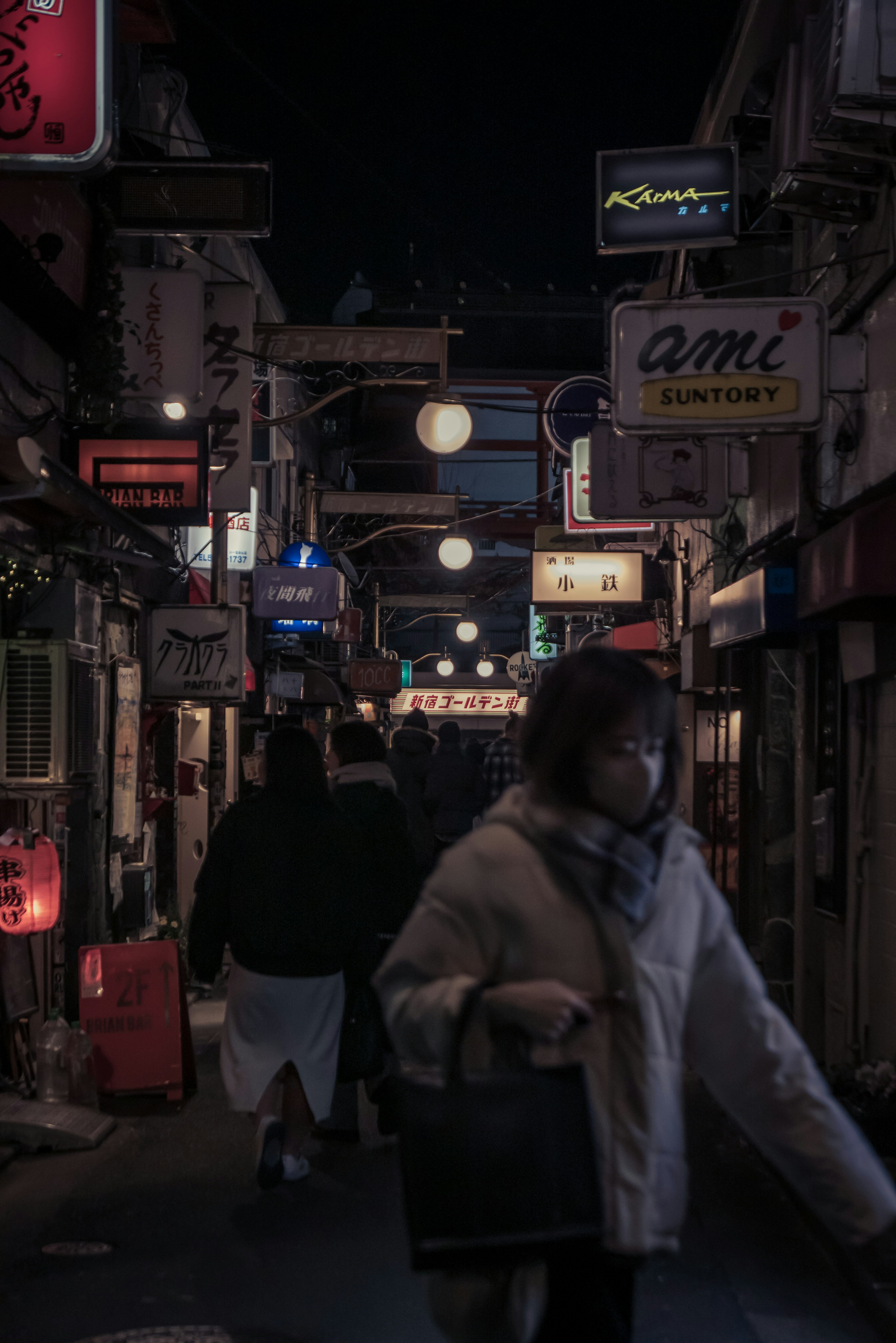 Imagen de una calle poco iluminada con personas caminando frente a tiendas con letreros de neón y faroles