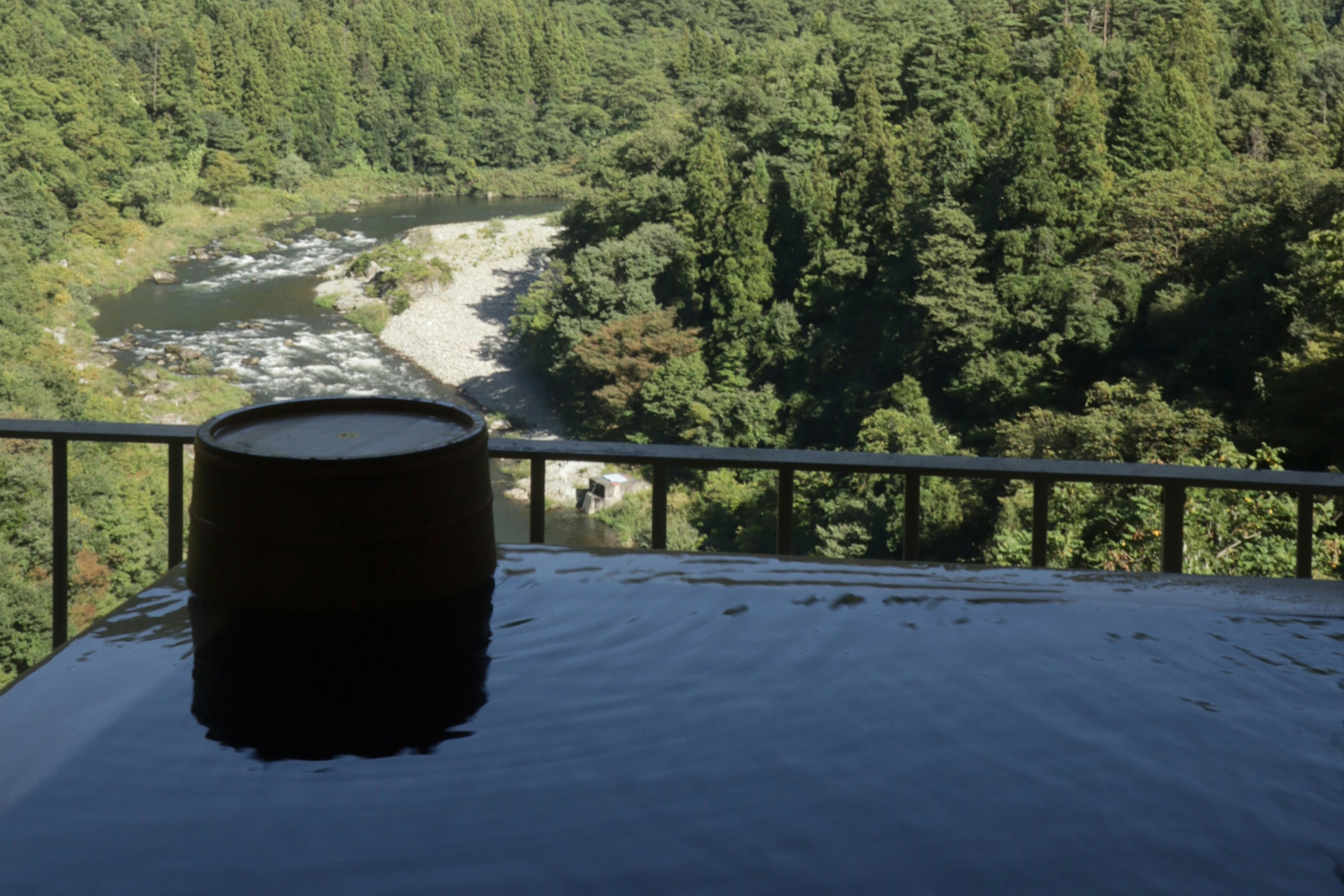 Scenic view of an outdoor hot spring with a wooden tub overlooking a river and lush greenery