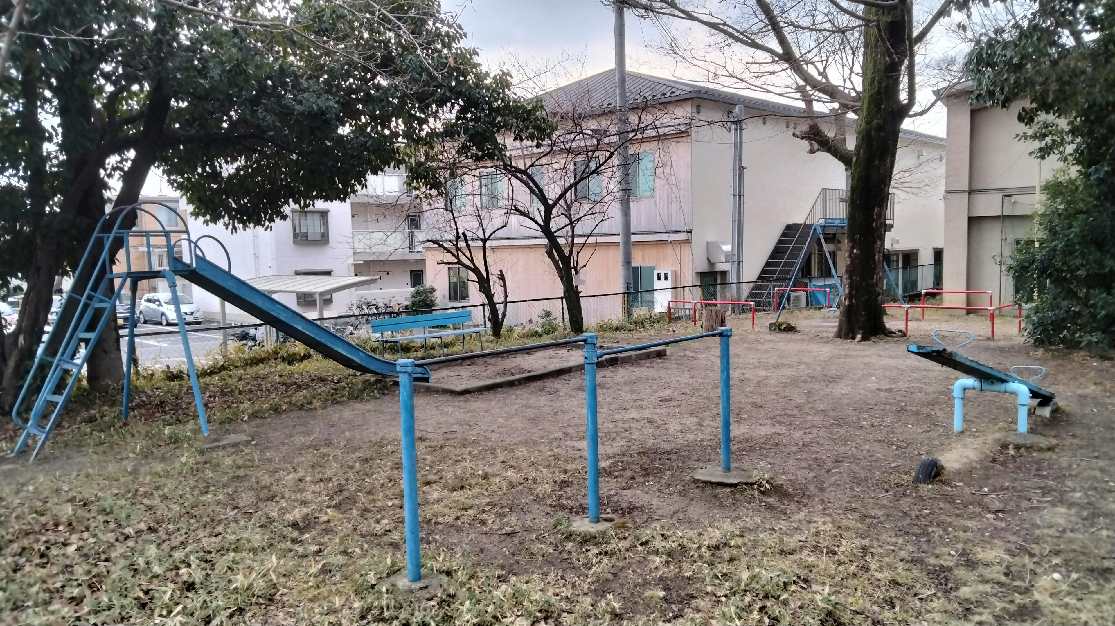 View of an old playground featuring a blue metal slide and swing surrounded by trees and buildings