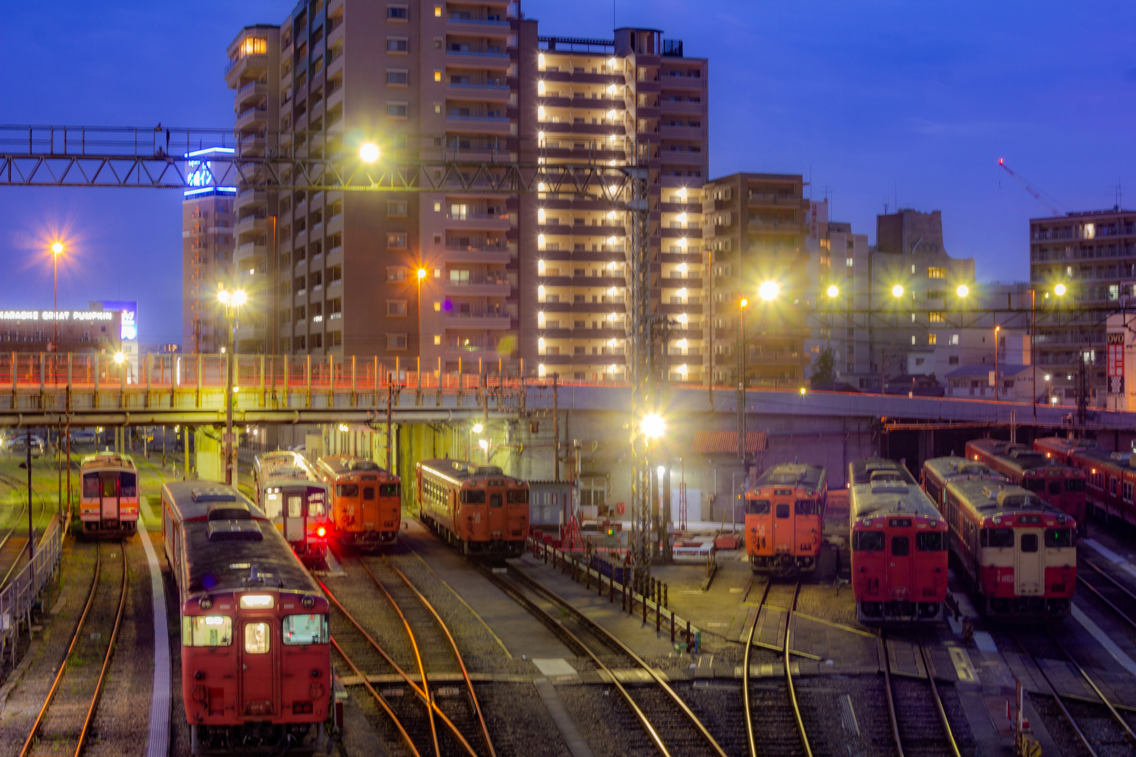 ทัศนียภาพสถานีรถไฟในเวลากลางคืน มีรถไฟหลายขบวนและอาคารสูง