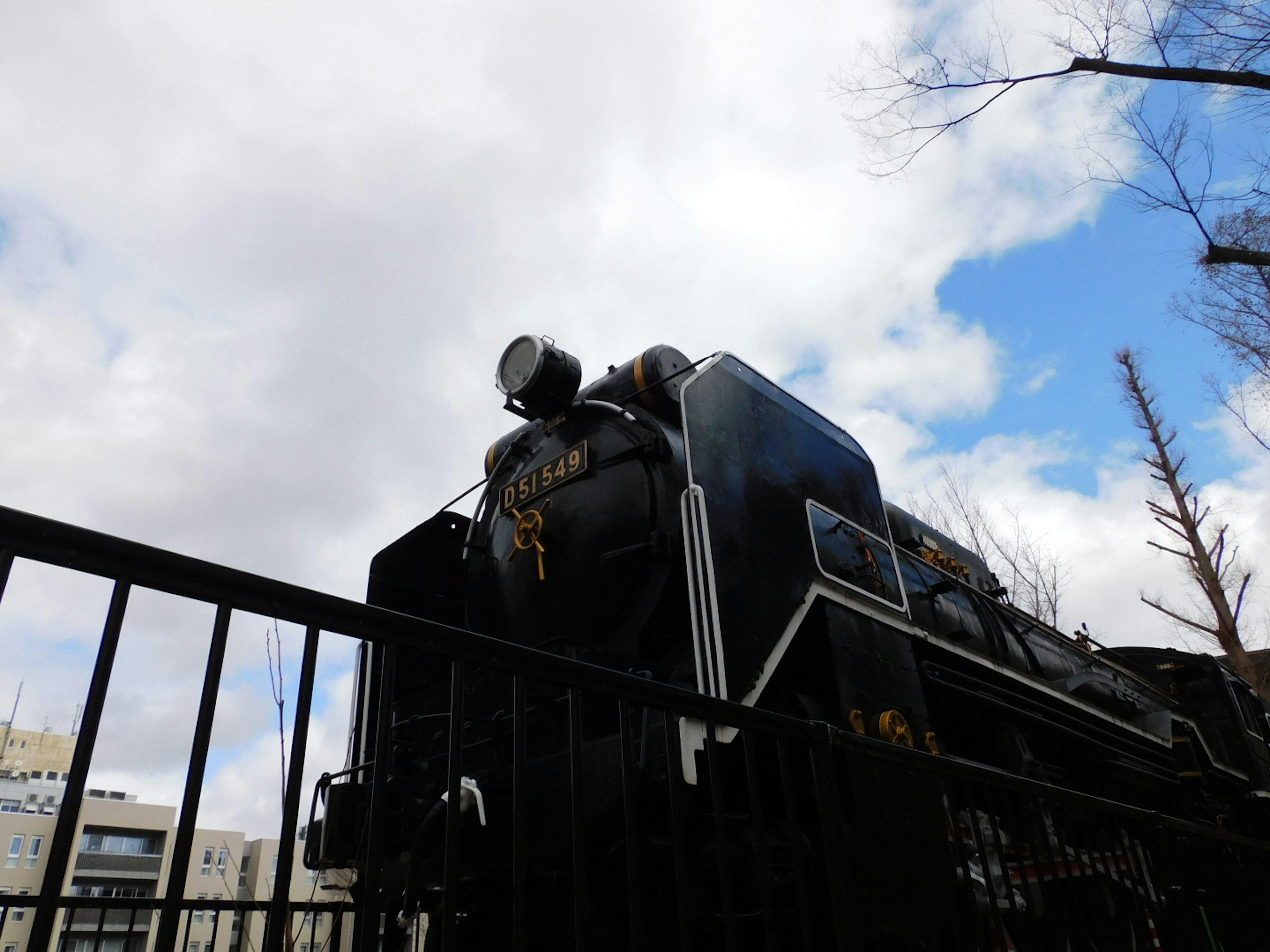 Black steam locomotive visible over a fence with blue sky and clouds in the background