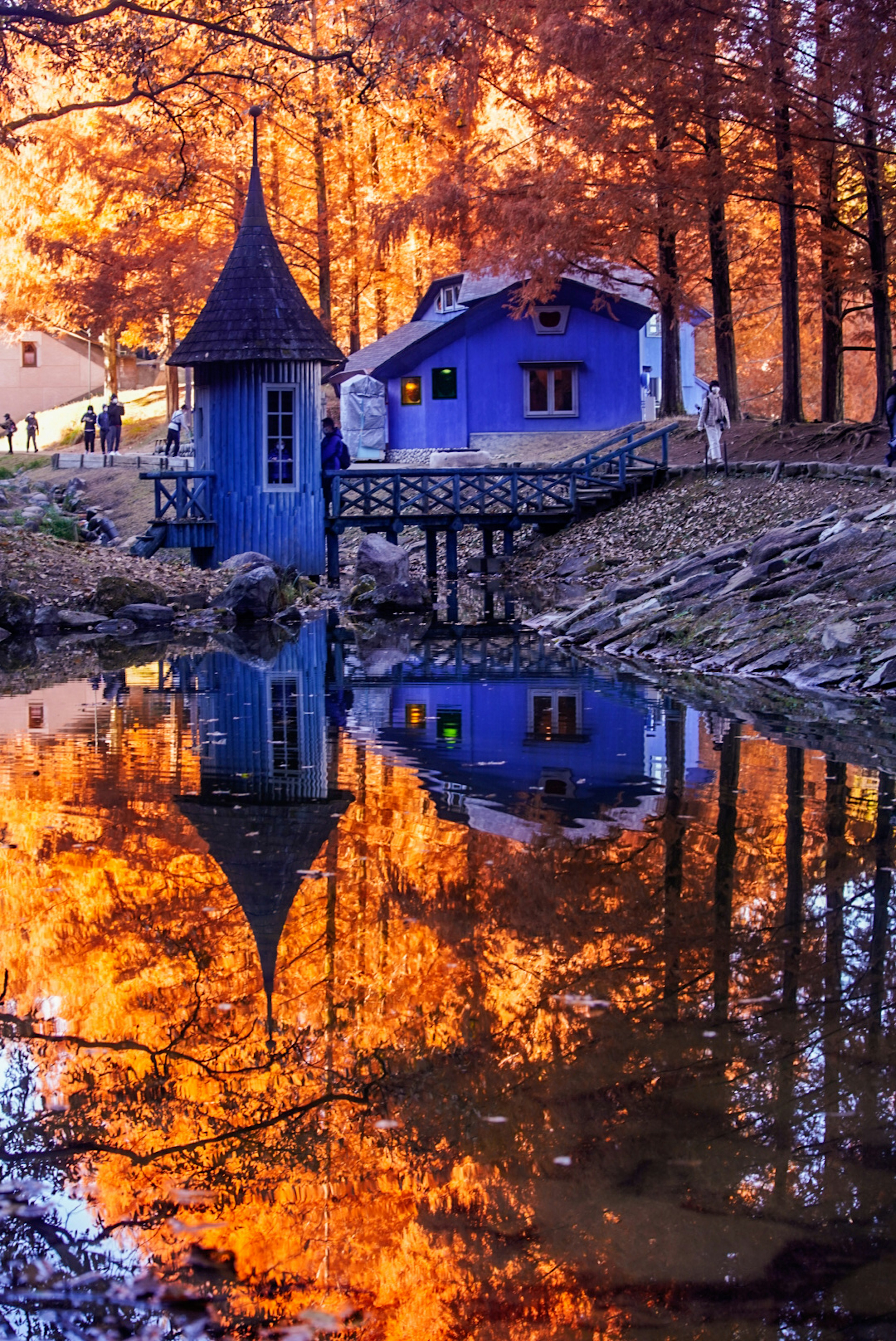 Eine ruhige Szene mit einer blauen Hütte und einer kleinen Brücke, die sich auf ruhigem Wasser spiegelt, umgeben von herbstlichem Laub