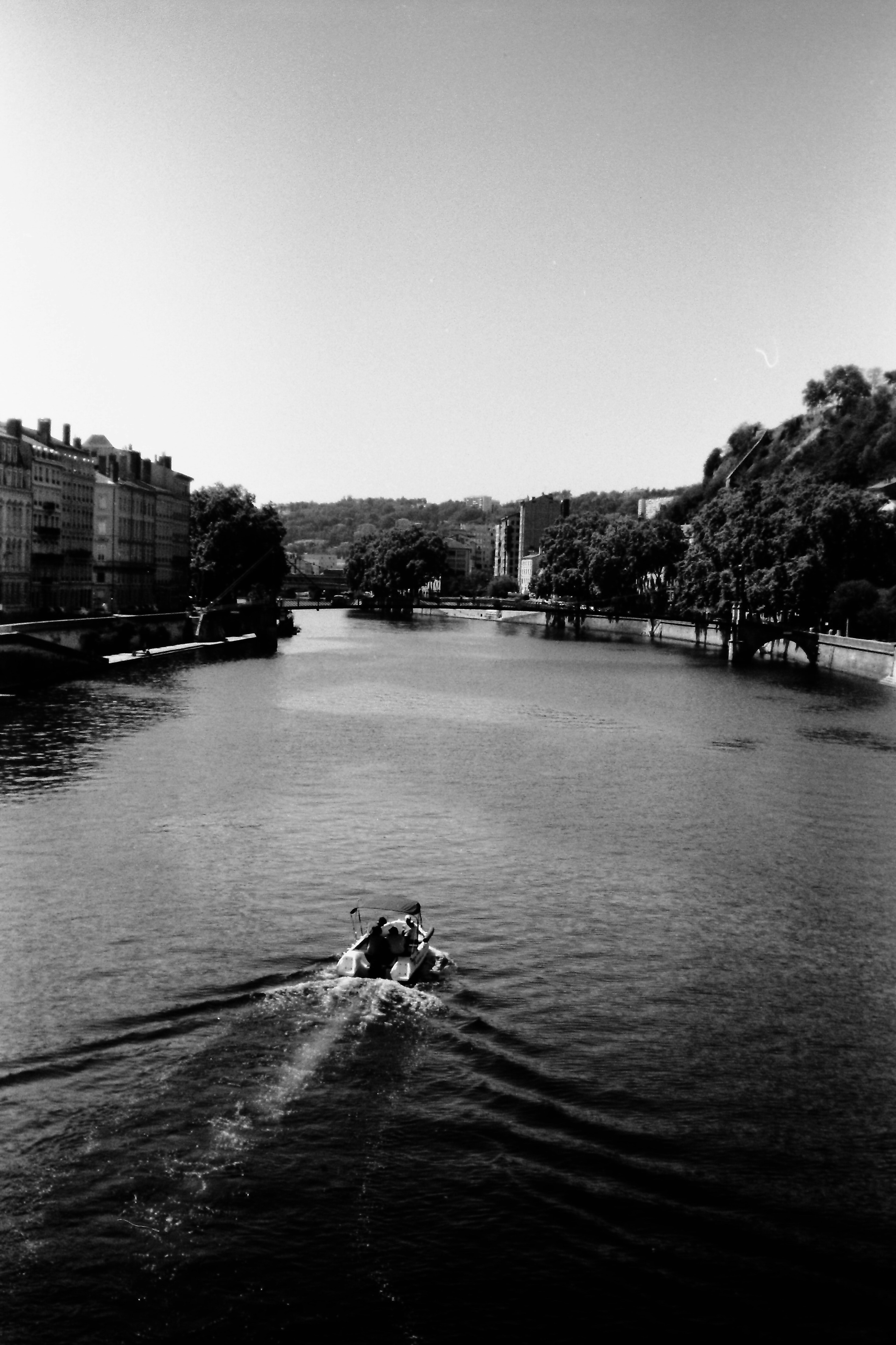 Black and white photo of a boat moving on a river surrounded by buildings and greenery