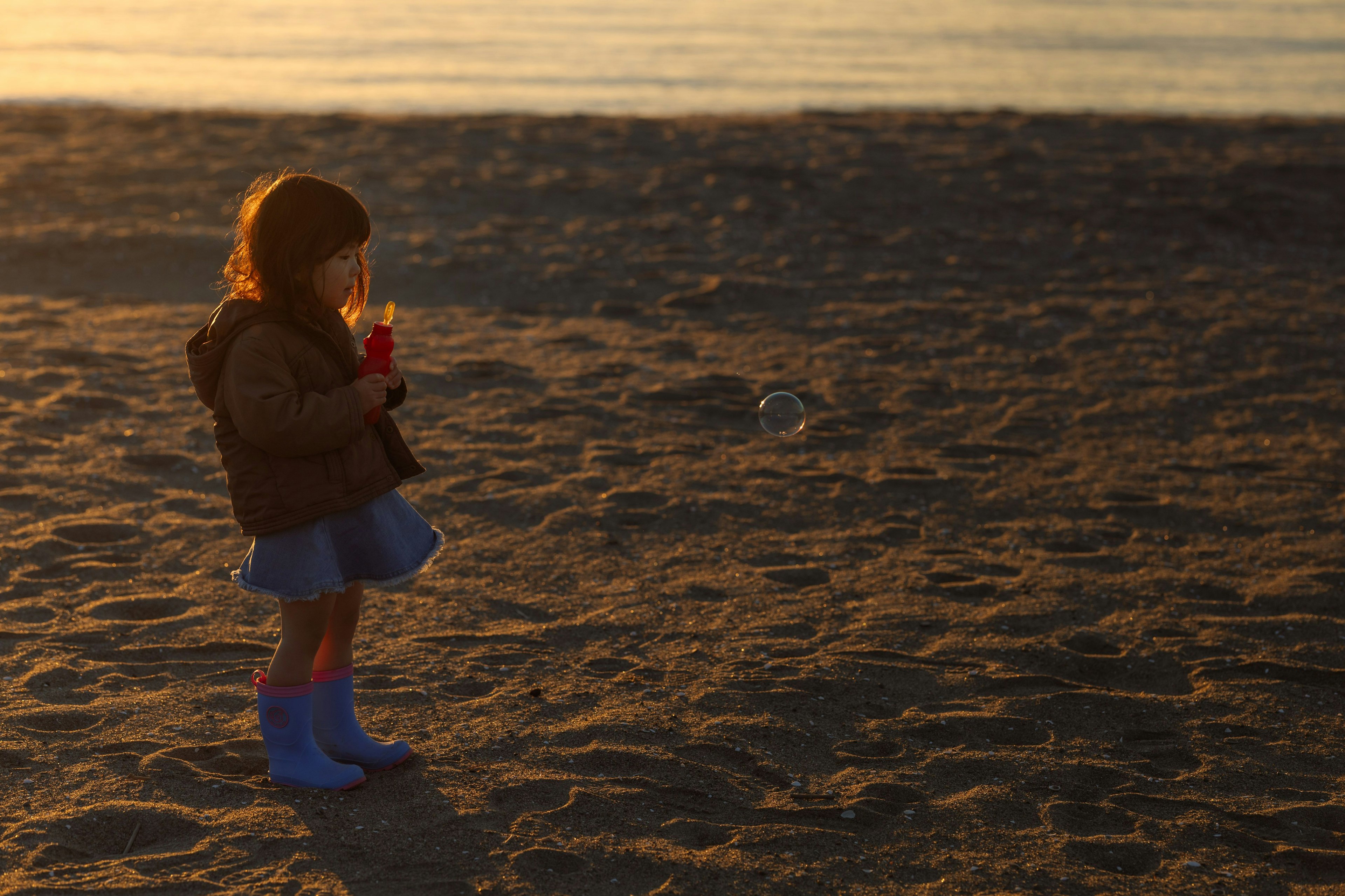 Kind steht am Sandstrand bei Sonnenuntergang hält ein rohes Getränk trägt blaue Stiefel