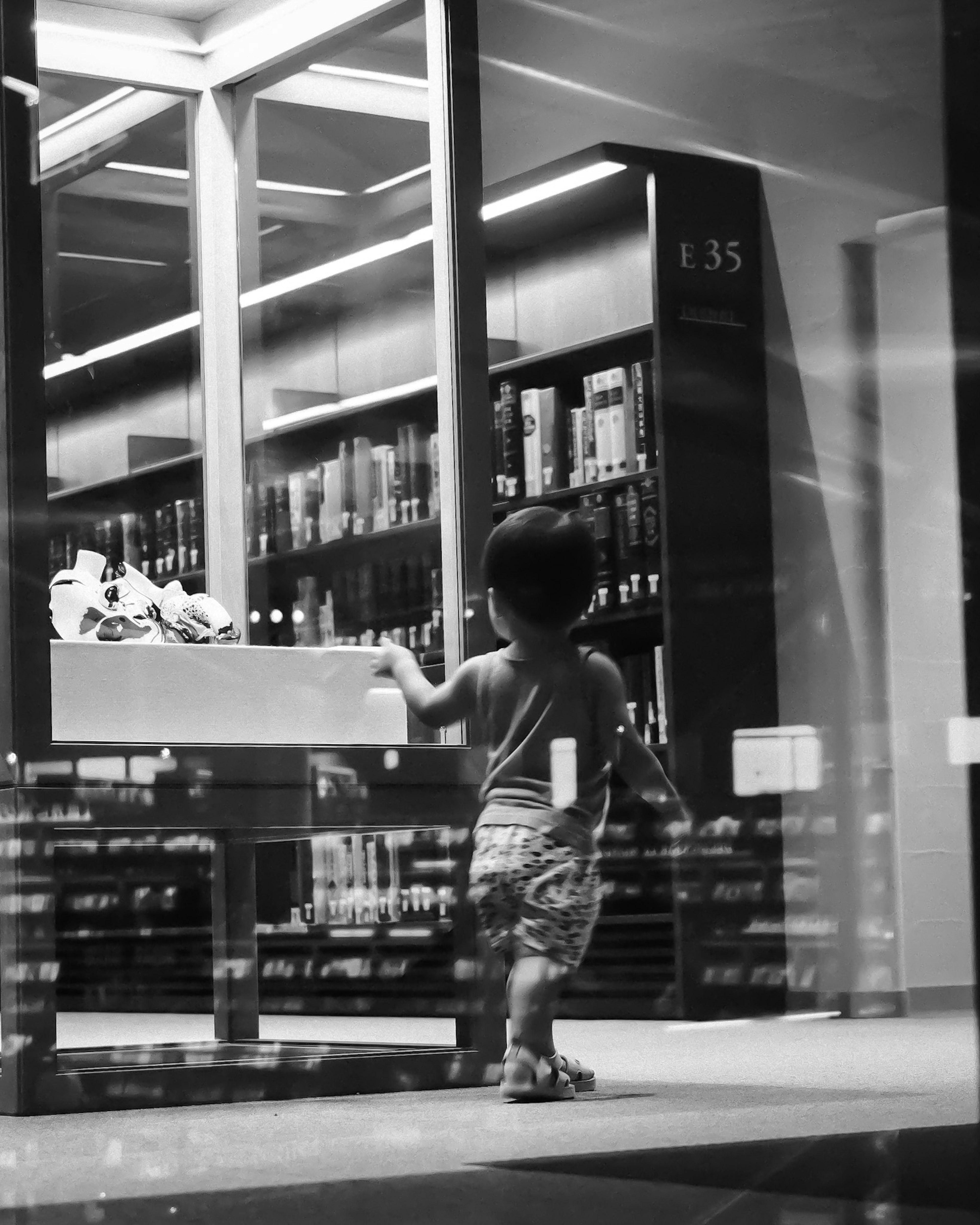 Un bambino piccolo in piedi davanti a uno scaffale di biblioteca con libri in bianco e nero