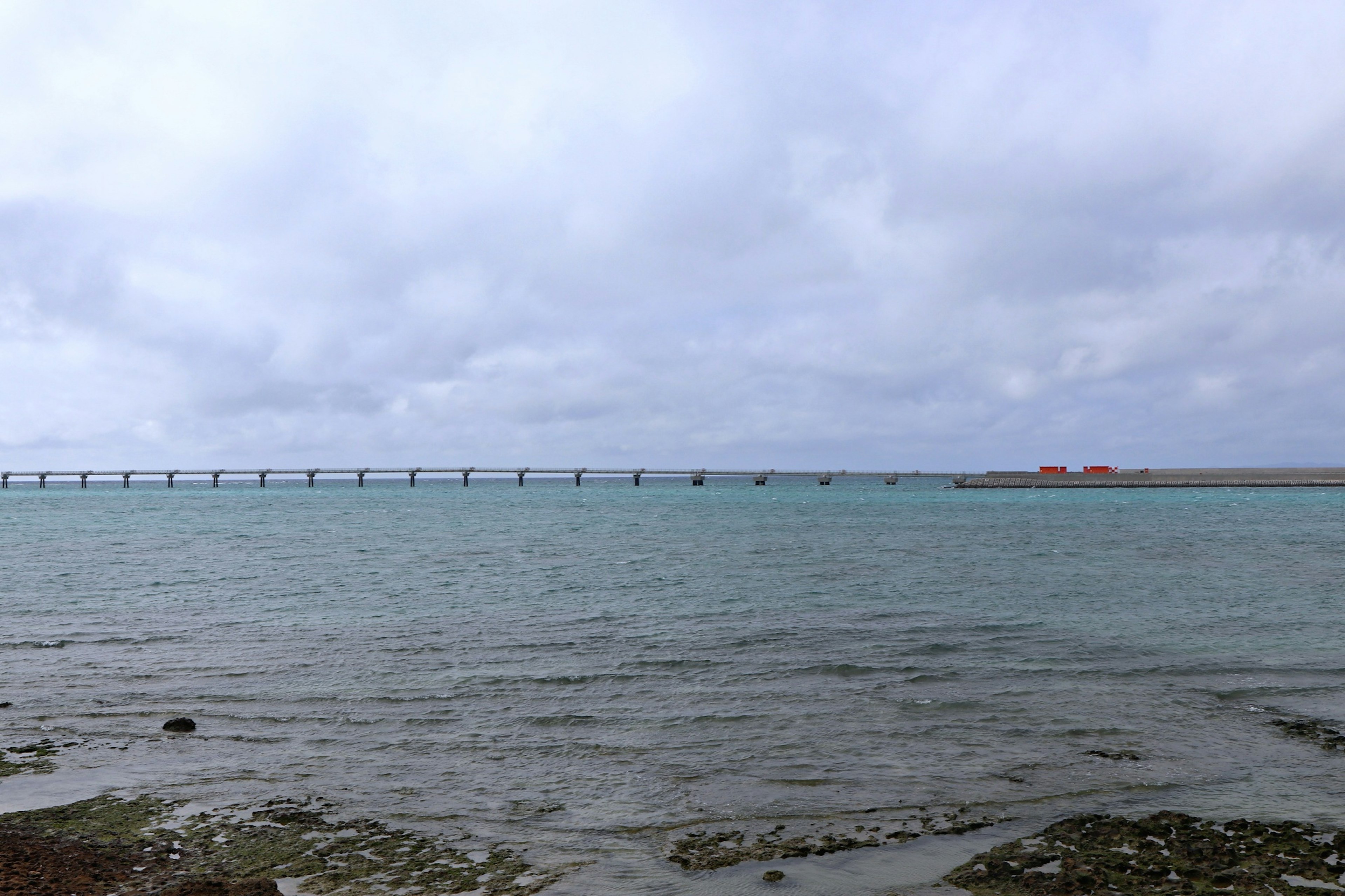 Lanskap yang menampilkan laut biru dan langit mendung dengan jembatan di kejauhan