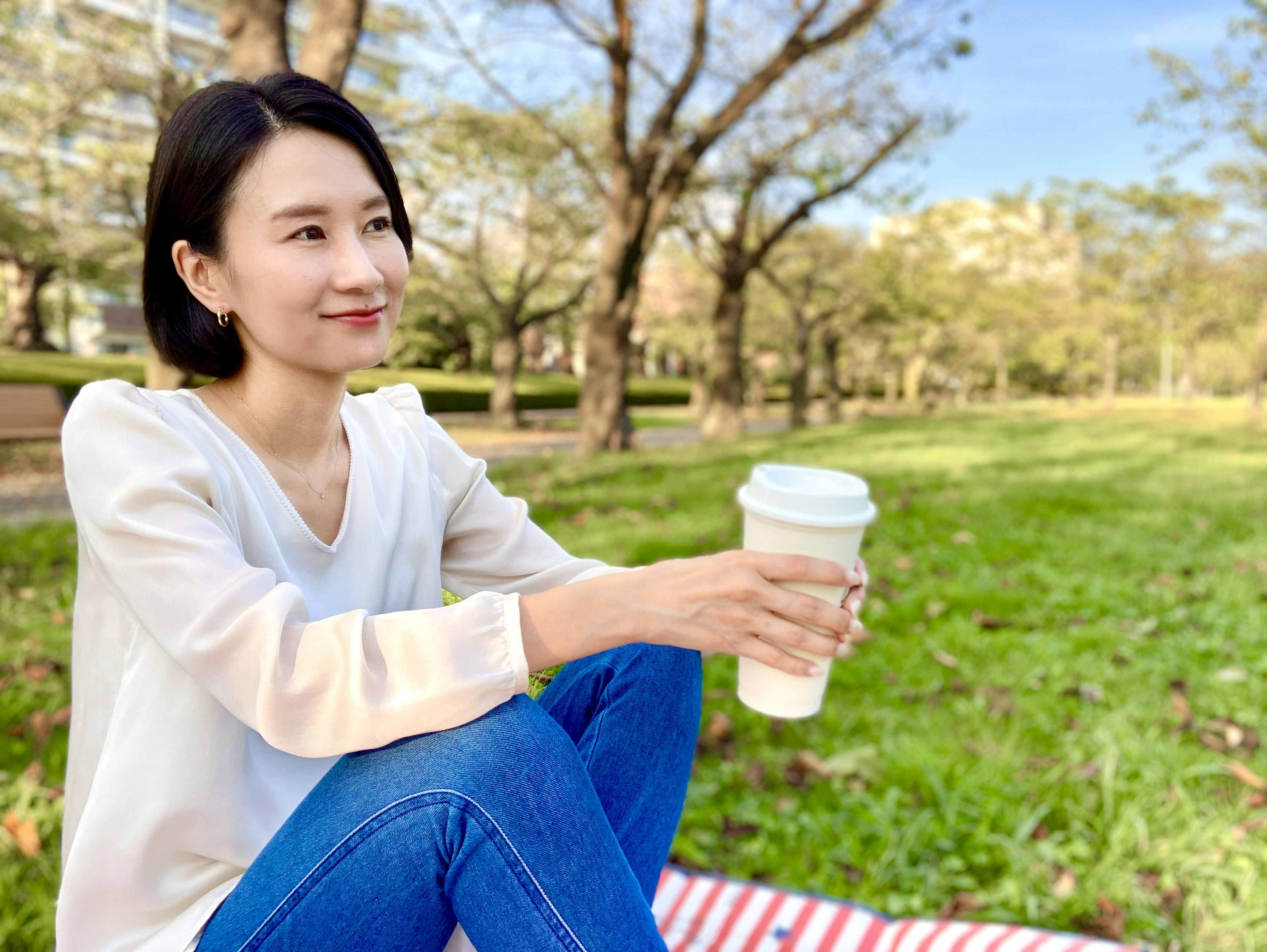 Frau mit Kaffeebecher, die auf dem Gras in einem Park sitzt, mit einem ruhigen Ausdruck und Bäumen im Hintergrund