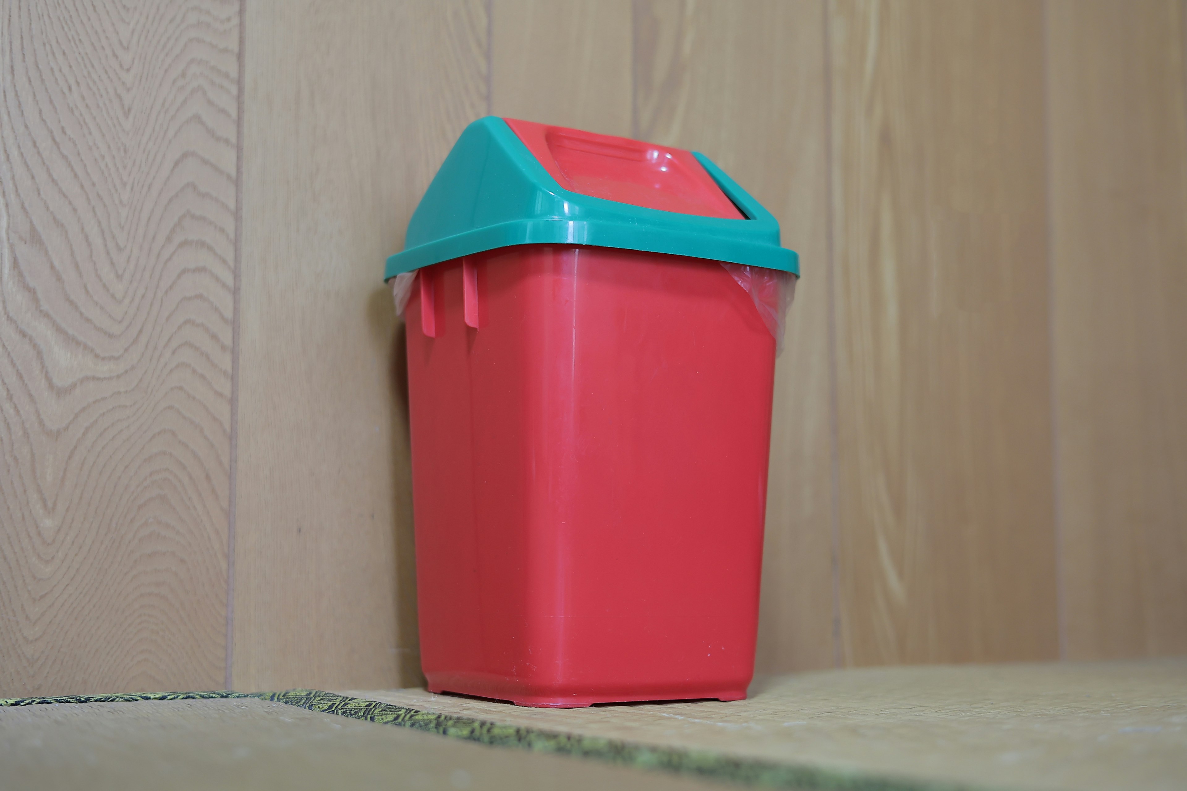 A red trash can with a blue lid leaning against a wall