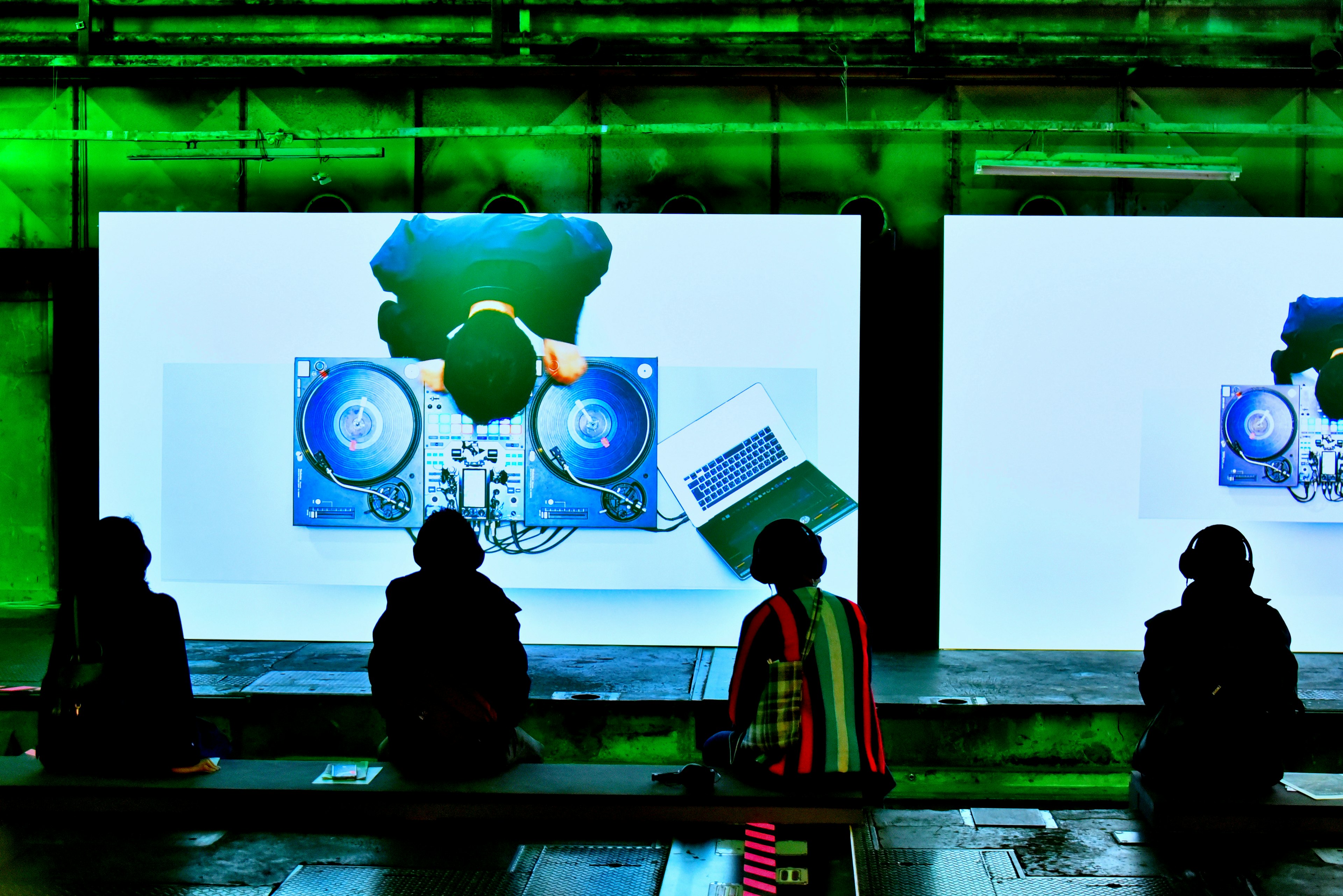 Personas sentadas frente a grandes pantallas que muestran equipo de DJ bajo una iluminación verde