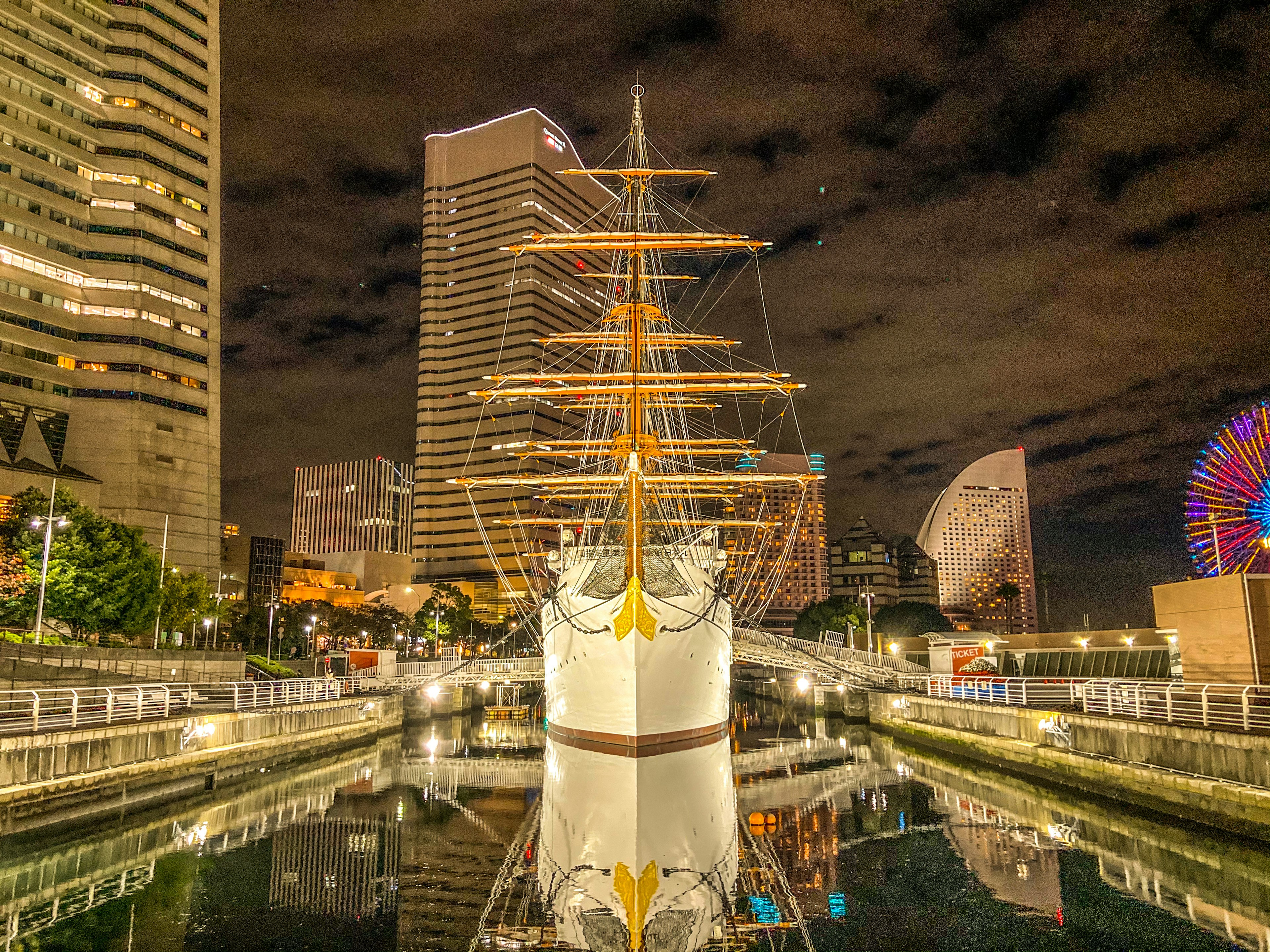 A tall ship illuminated at night in Yokohama harbor with modern buildings in the background