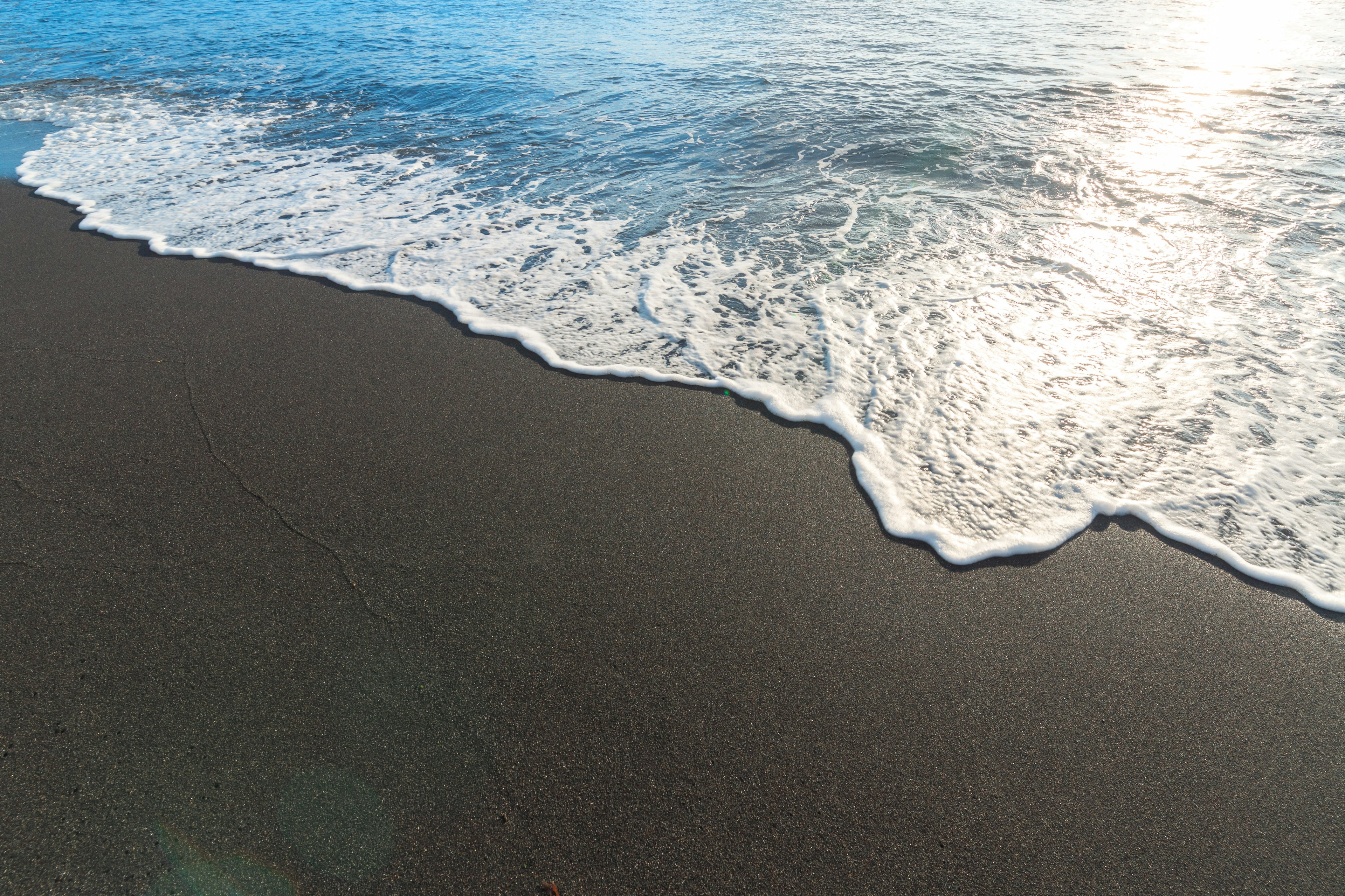 Wellen, die sanft über einen dunklen Sandstrand mit einem blauen Ozean rollen