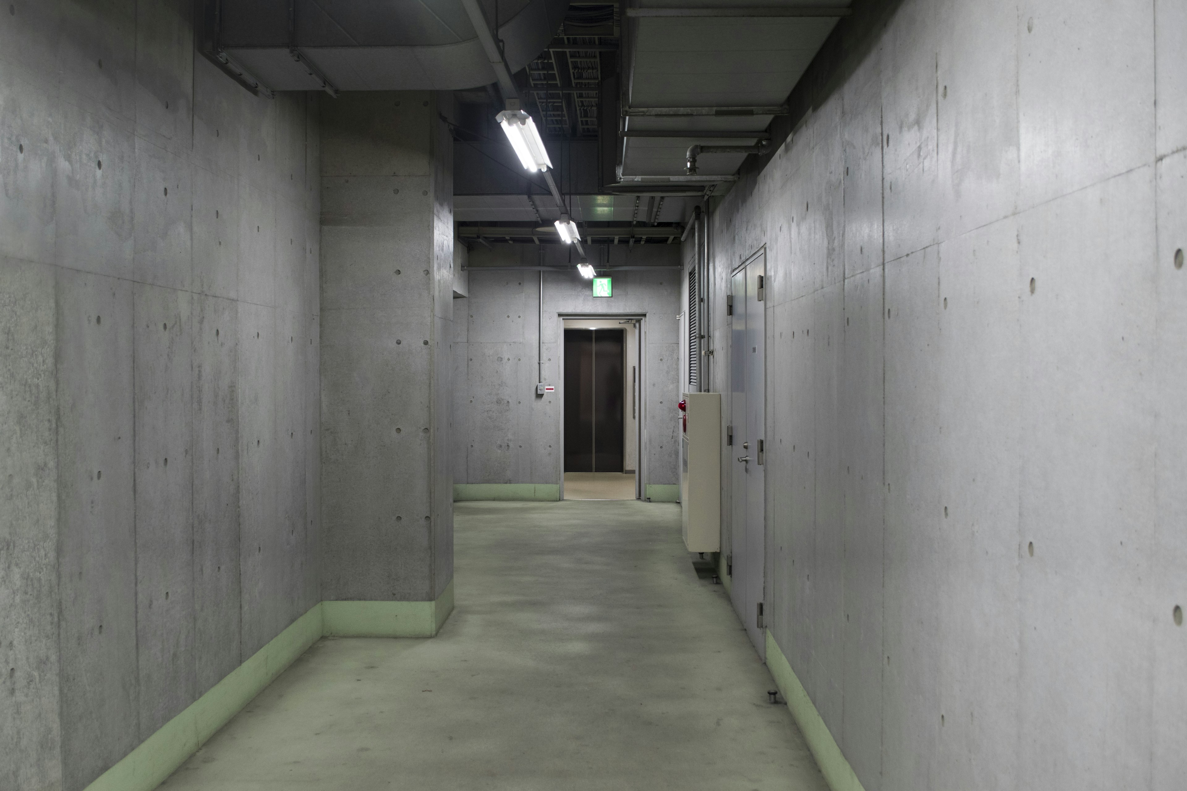 Long corridor with concrete walls and floor Ceiling lights illuminate the space A wooden door is visible at the end of the corridor