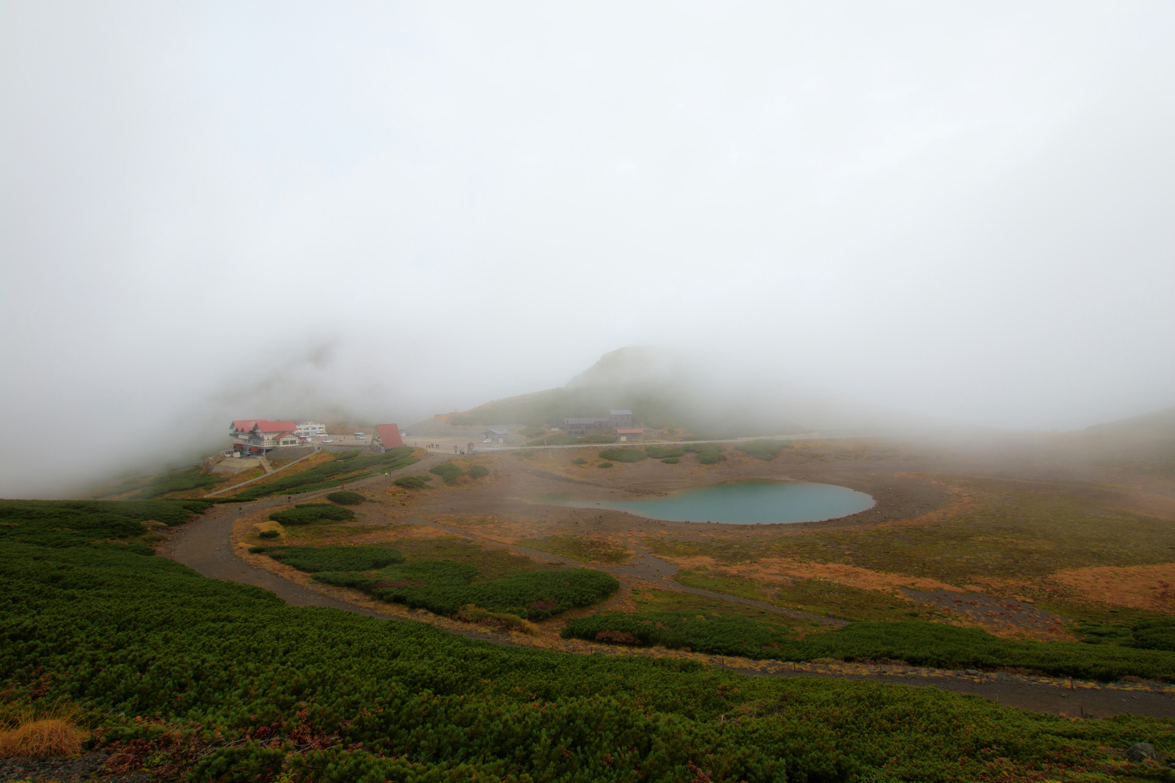 Paisaje montañoso con niebla que incluye un pequeño estanque y un edificio