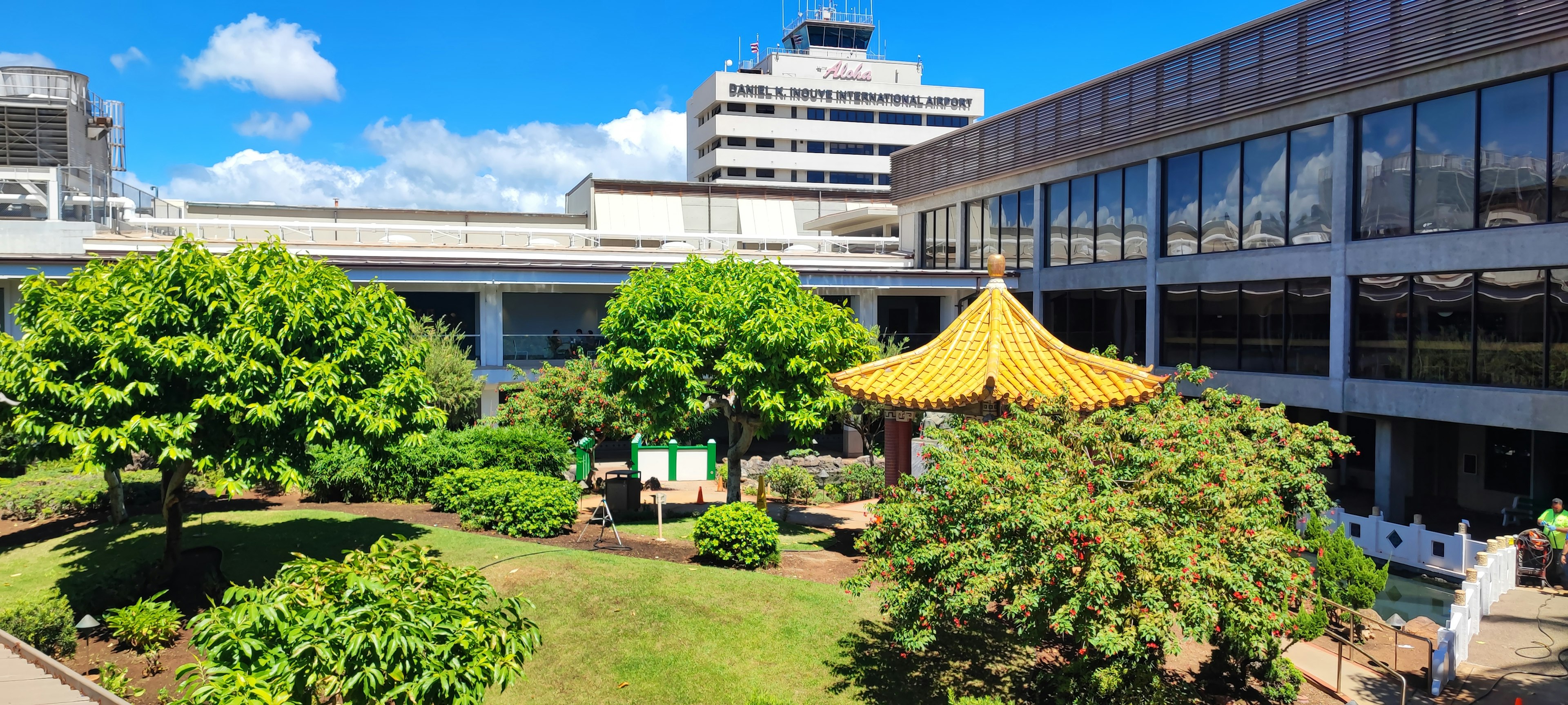 Jardín exuberante con un pabellón de techo amarillo y edificios modernos al fondo