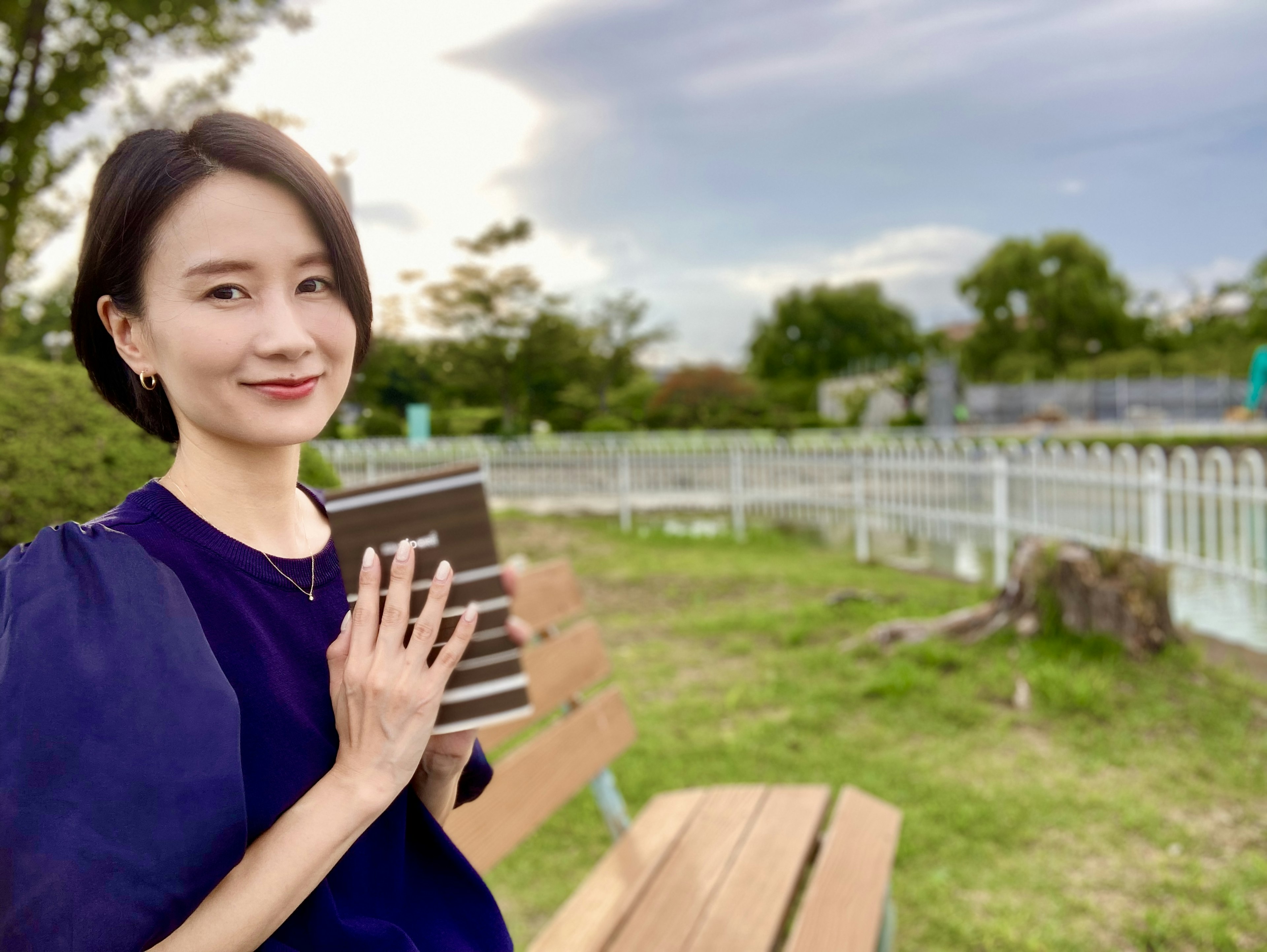 Eine Frau sitzt auf einer Parkbank und hält ein Buch in der Hand und lächelt