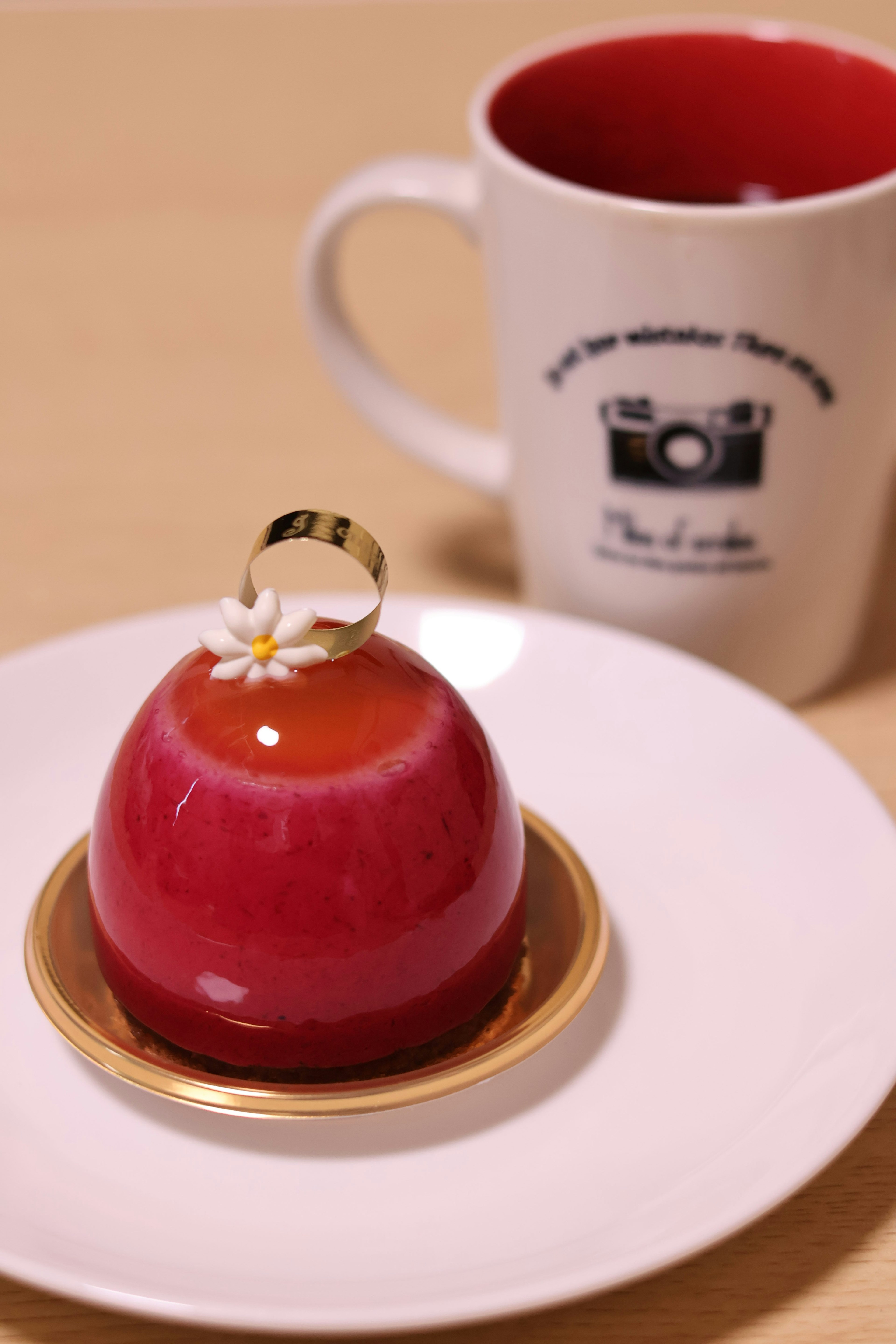 Un hermoso postre rojo en un plato blanco junto a una taza con diseño de cámara