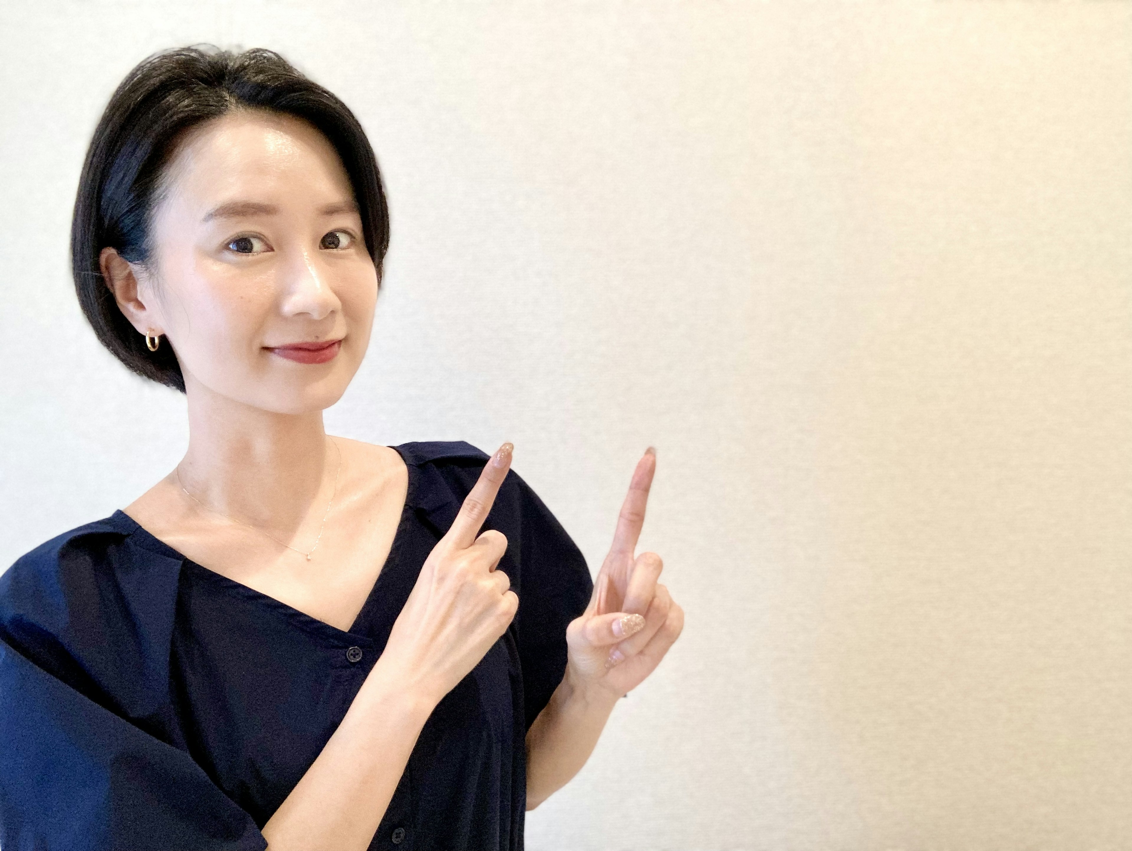 A woman smiling and pointing with both hands against a simple cream-colored wall