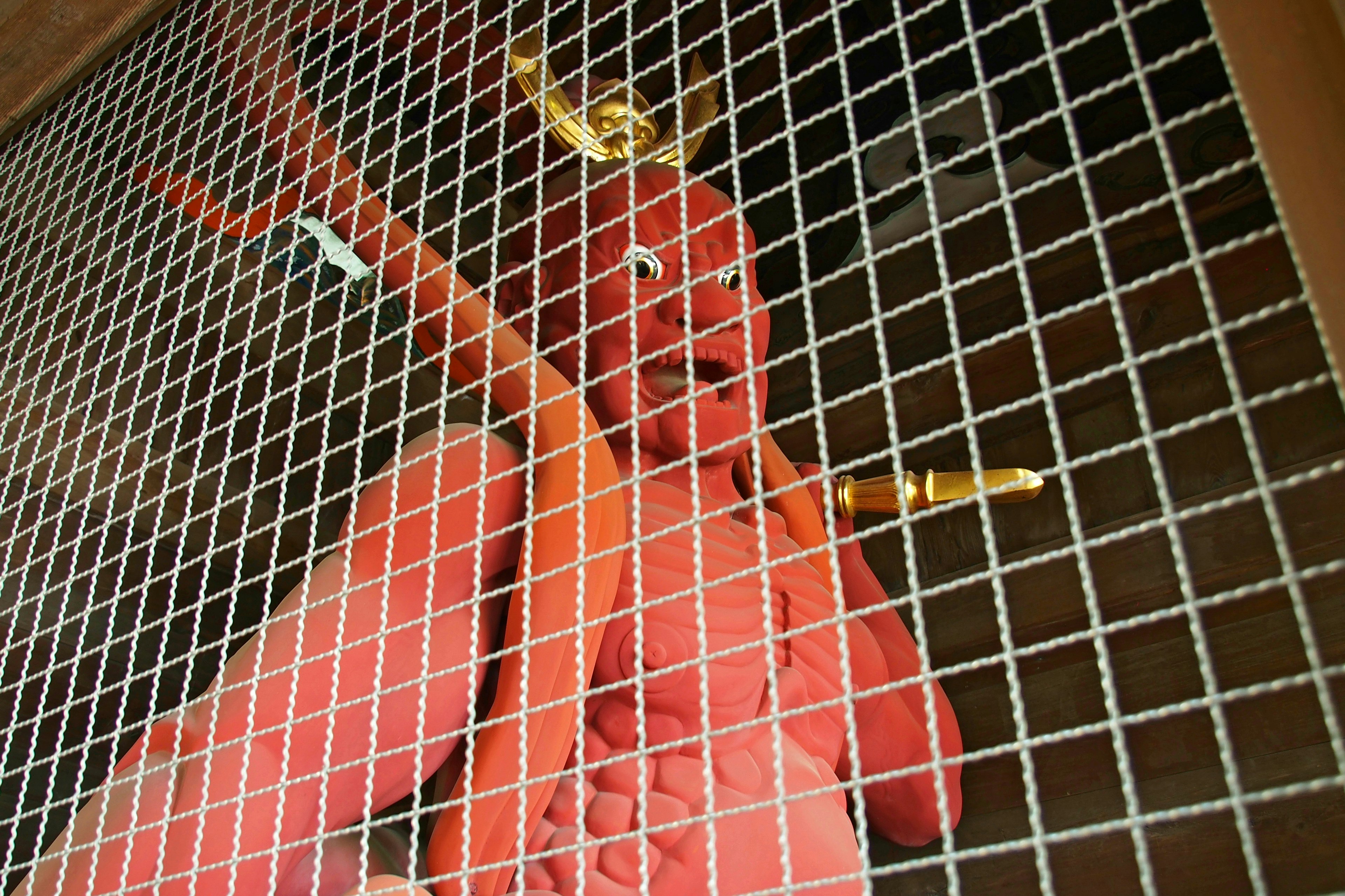 A red demon statue wearing a golden crown behind a metal grid