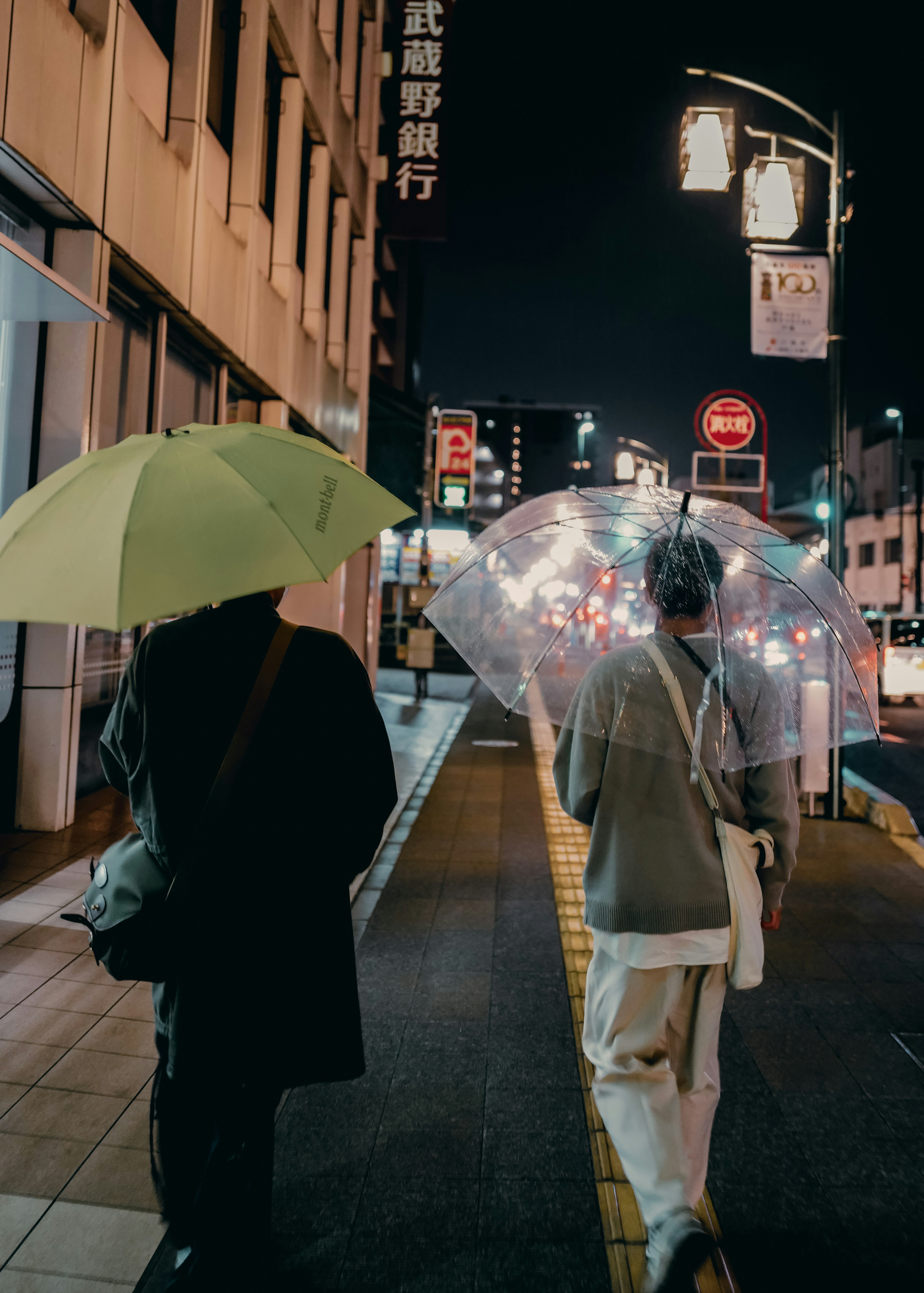 夜の街を歩く二人の人物 一人は緑の傘を持ち もう一人は透明な傘を持つ