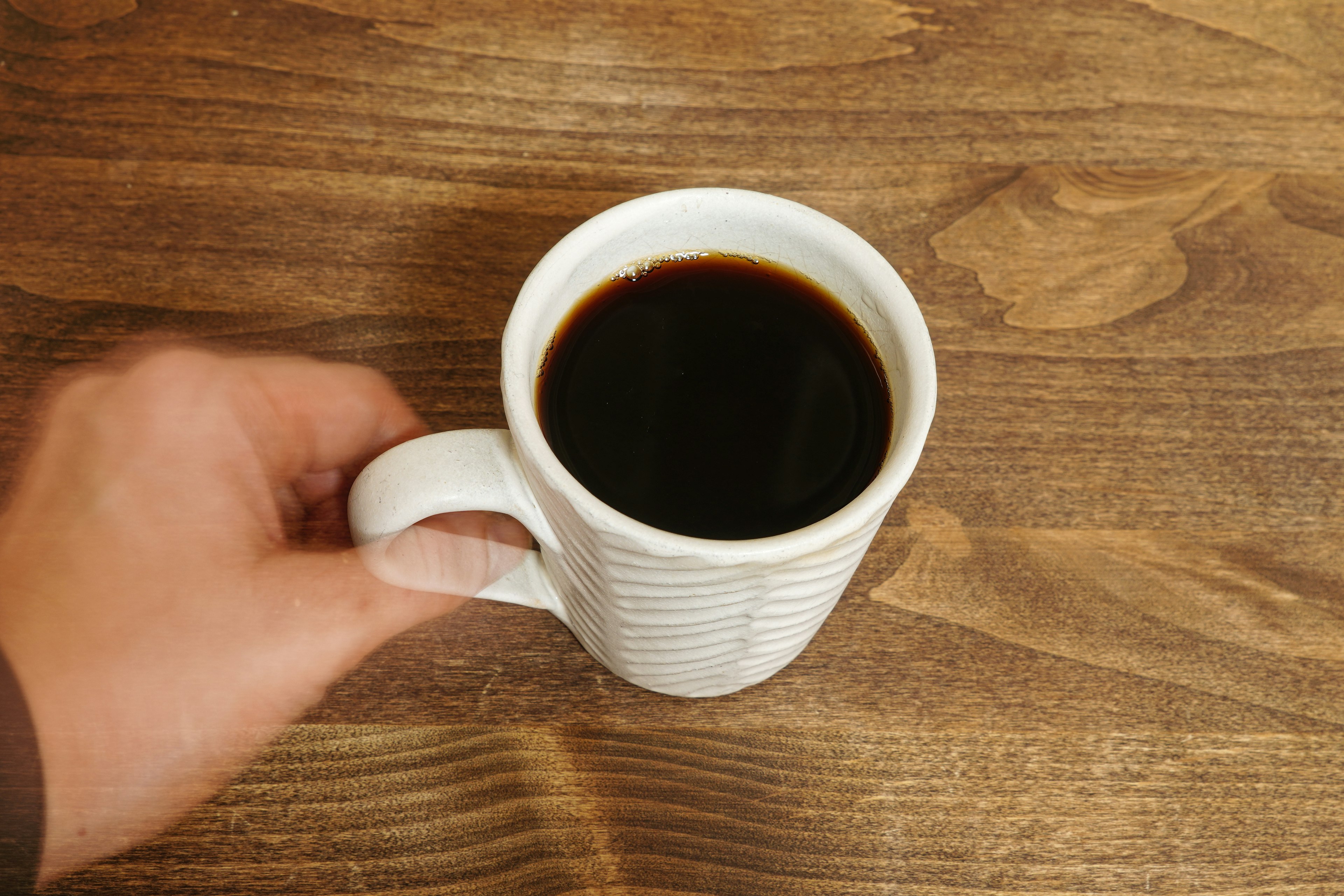 A hand holding a white coffee cup filled with black coffee on a wooden table