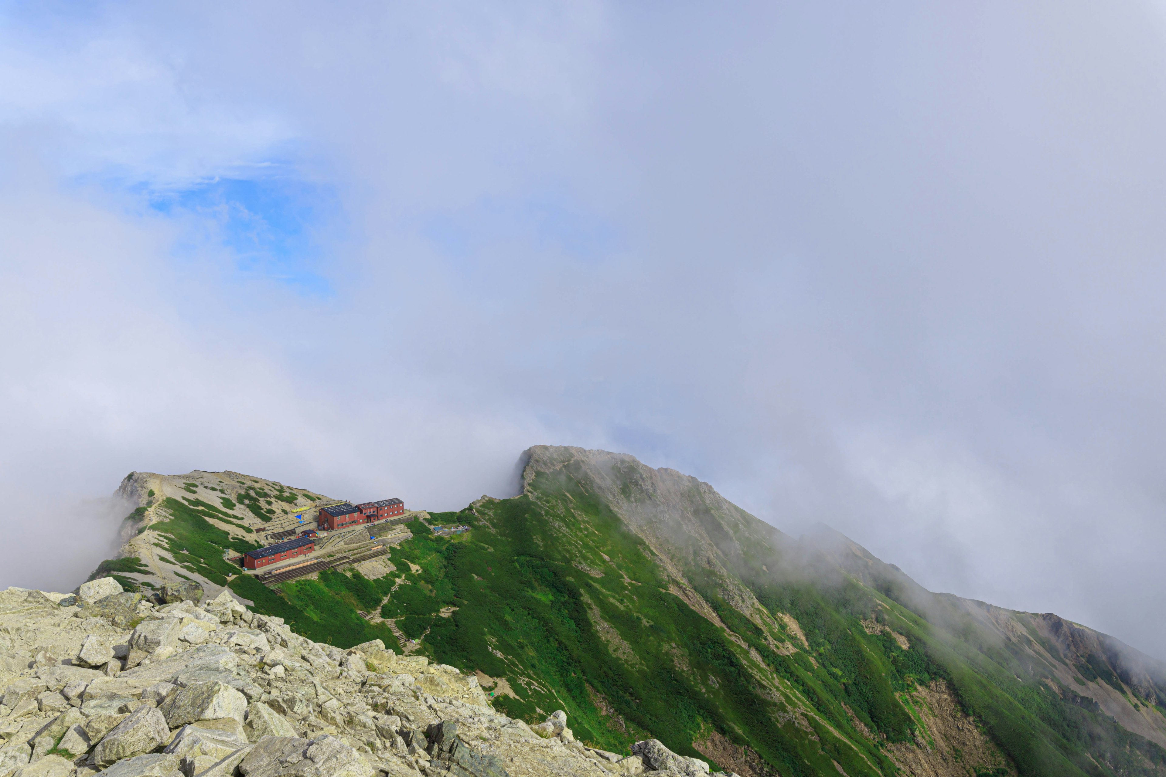 Paysage montagneux avec un cabanon sur une pente verte partiellement couvert par des nuages