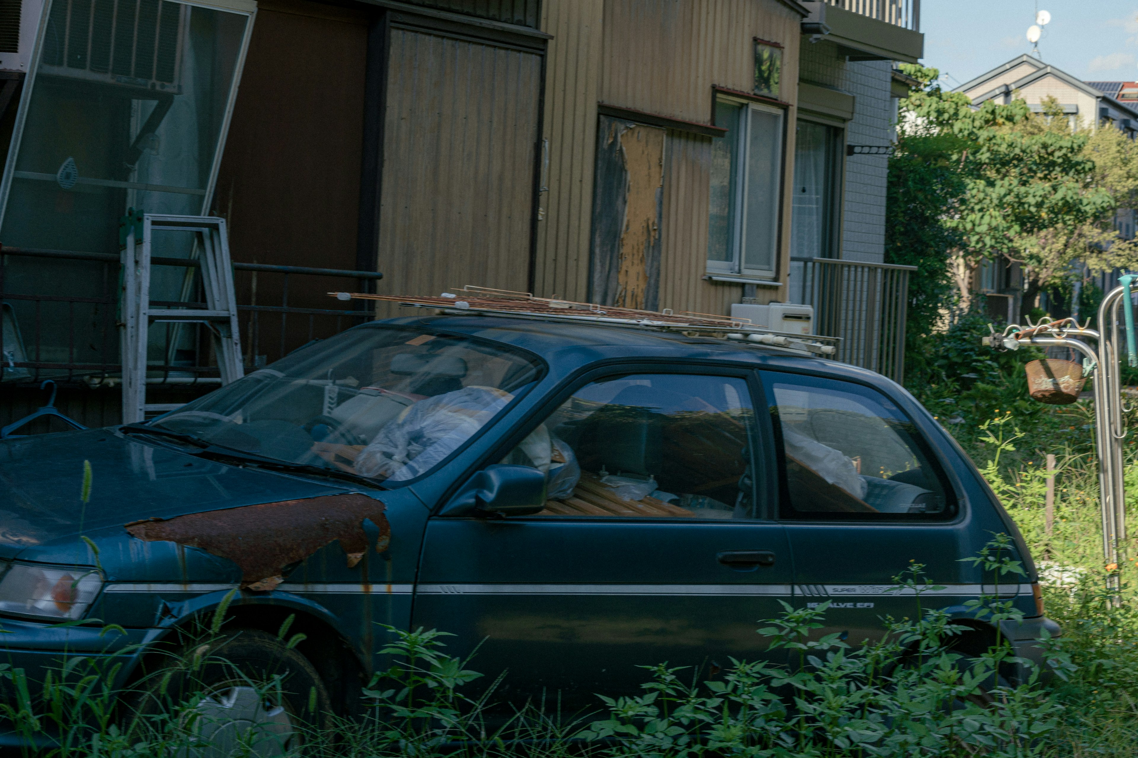 An old blue car parked next to a dilapidated house overgrown with weeds