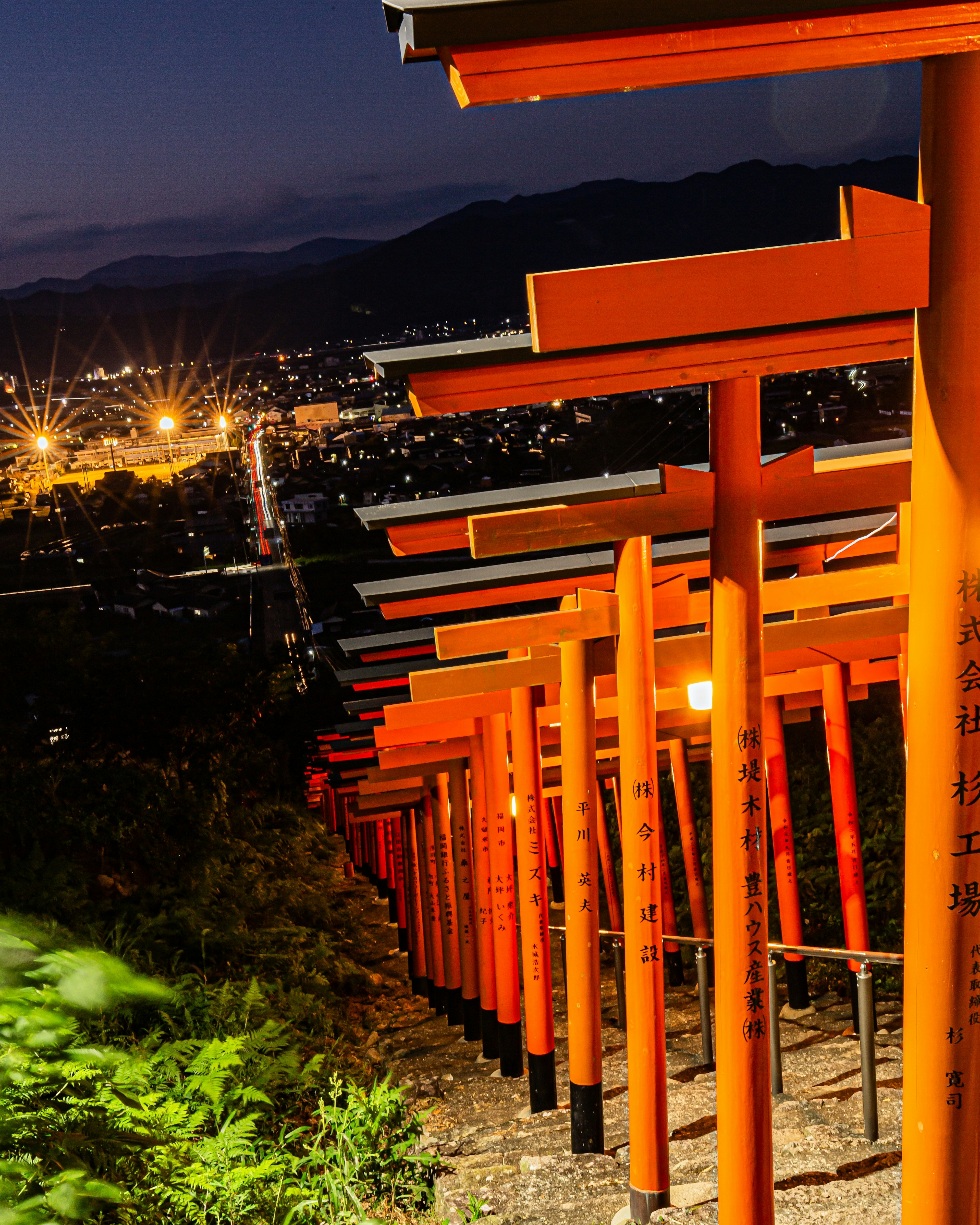 Vibrant red torii gates lining a mountain path at night with city lights in the distance